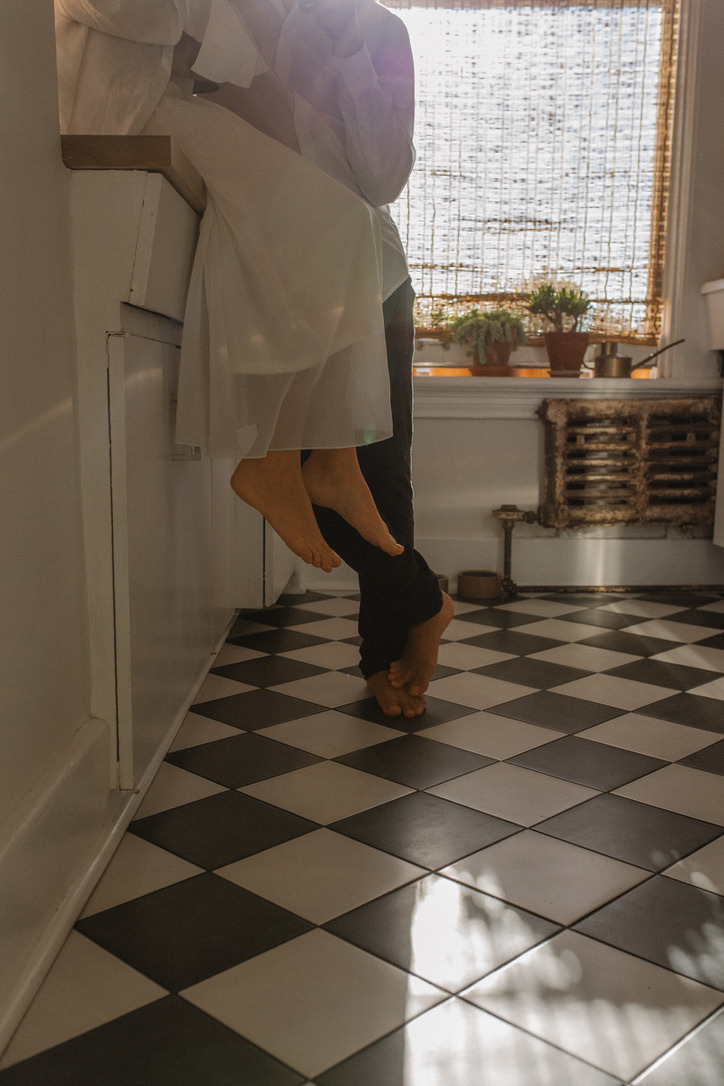 A person wearing a white dress and another person in dark pants are hanging upside down with their heads near the floor and feet near the ceiling, standing on a black and white checkered tile floor in a room with a window, potted plants, and a radiator.