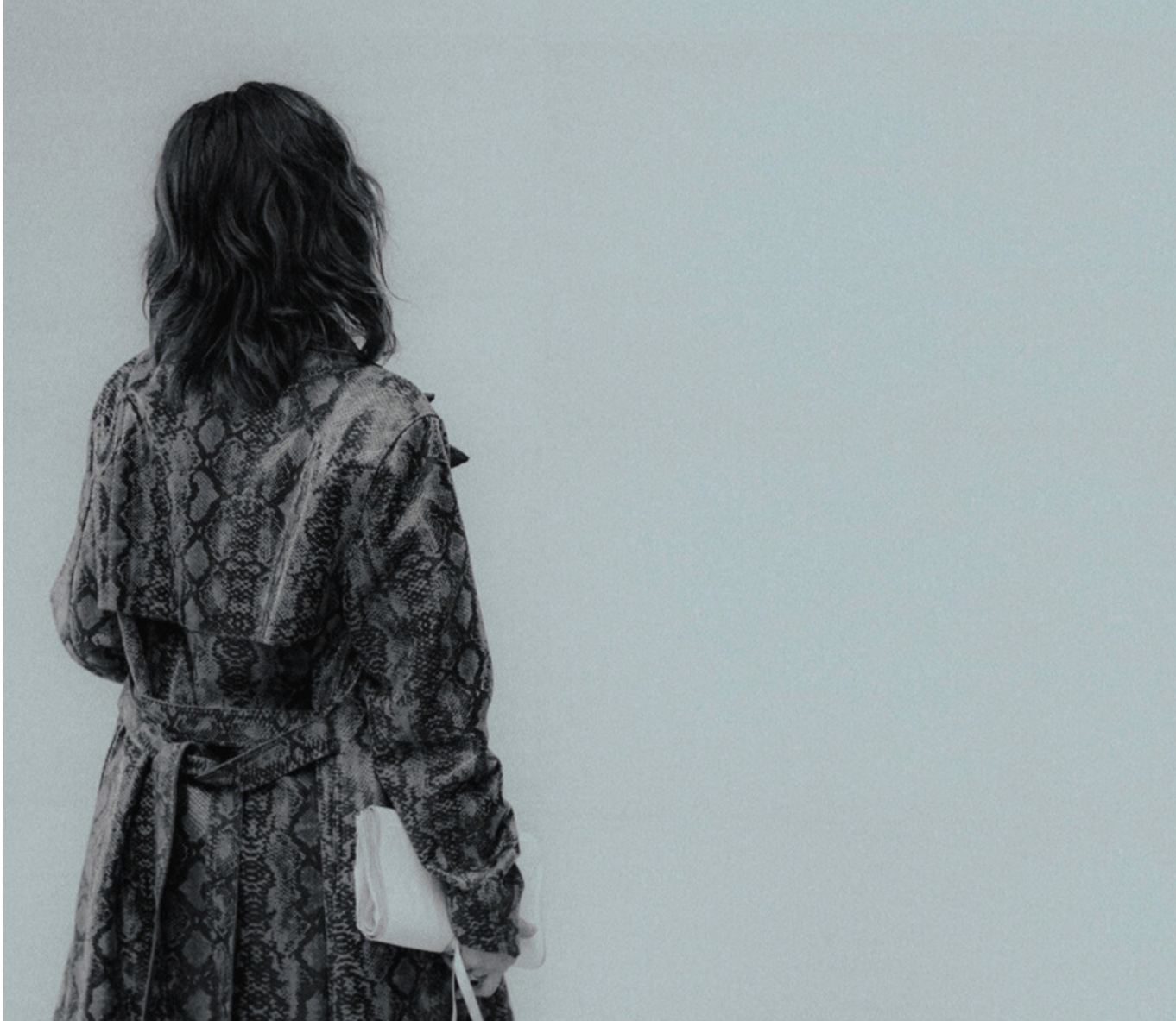A woman with medium wavy black hair wearing a snake print trench coat holding a white clutch bag, standing against a plain light grey wall.