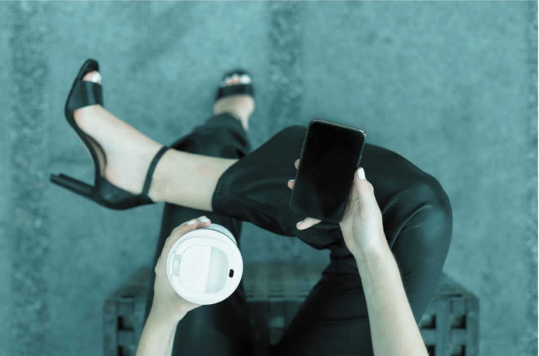 Woman sitting on a black crate, holding a smartphone in one hand and a coffee cup in the other, outdoors on a sidewalk.