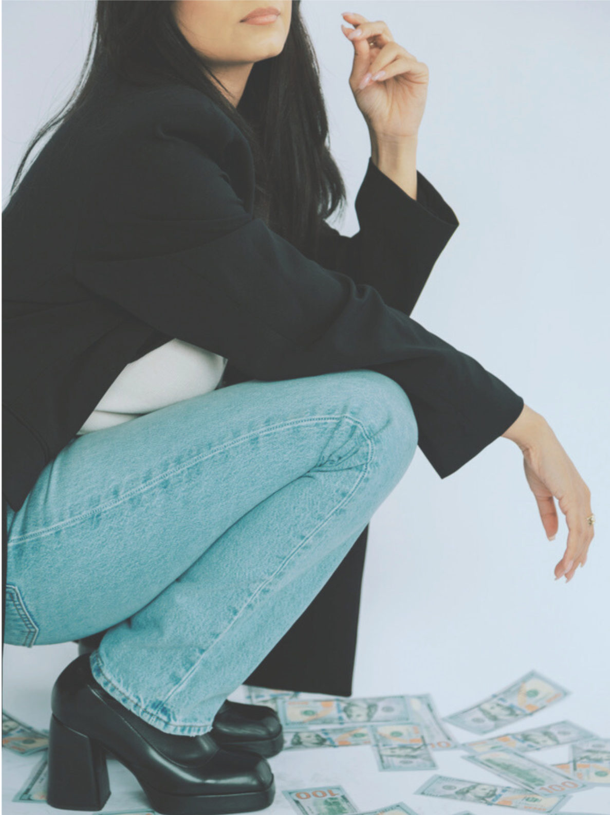 A woman with long dark hair, wearing a black blazer, white top, light blue jeans, and black platform shoes, squatting on the ground surrounded by scattered U.S. hundred-dollar bills.