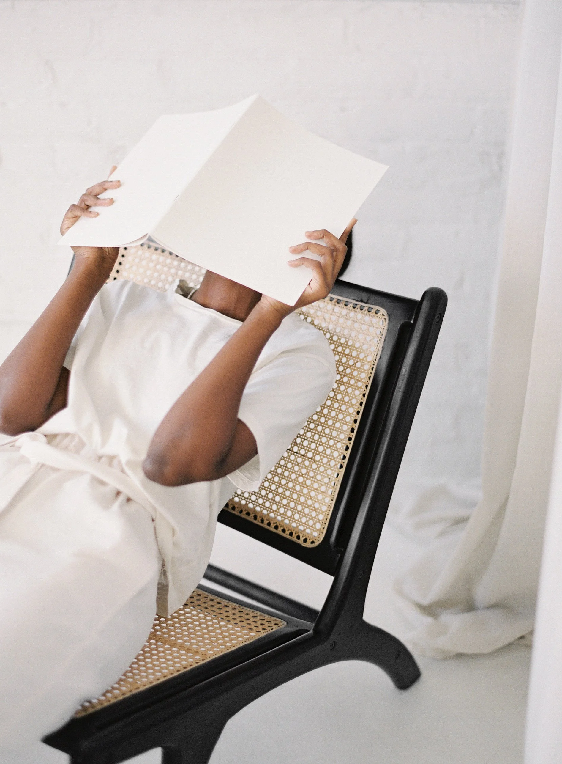 A person sitting on a chair with a woven cane seat and back, holding a white open book or notebook in front of their face, wearing a white dress, against a white wall background.