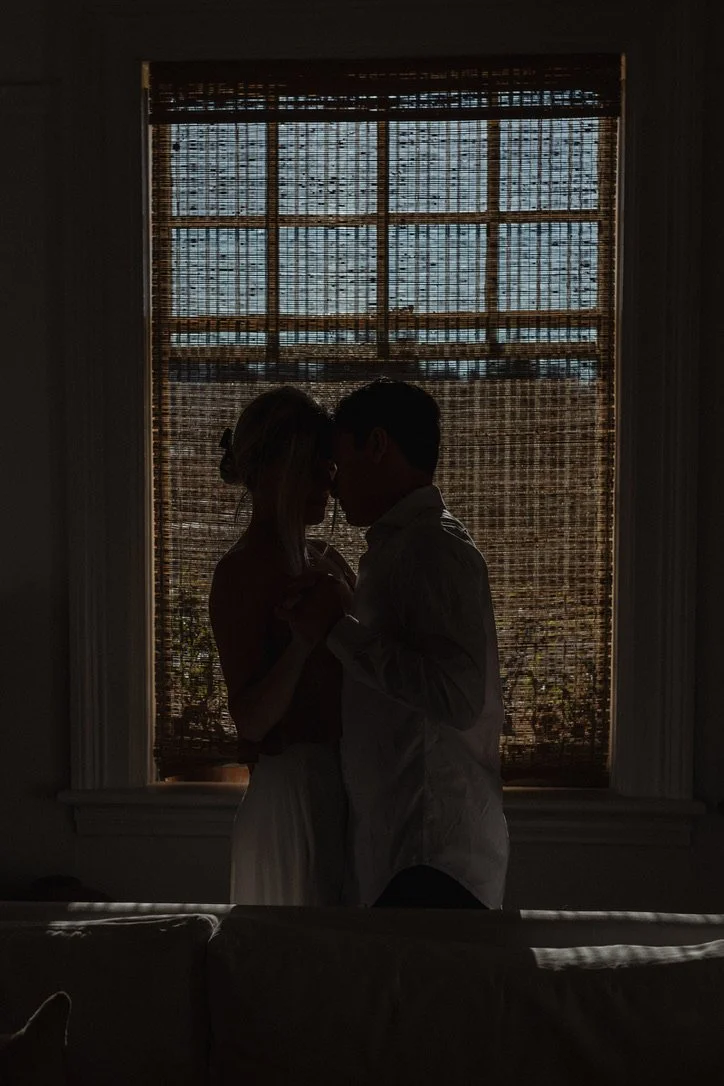 Silhouette of a couple standing close and holding hands in front of a window with woven blinds, backlit by sunlight.