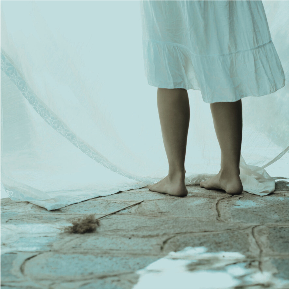 The lower half of a person standing barefoot on stone flooring in a light, flowing dress, with sheer curtains and a feather duster nearby.