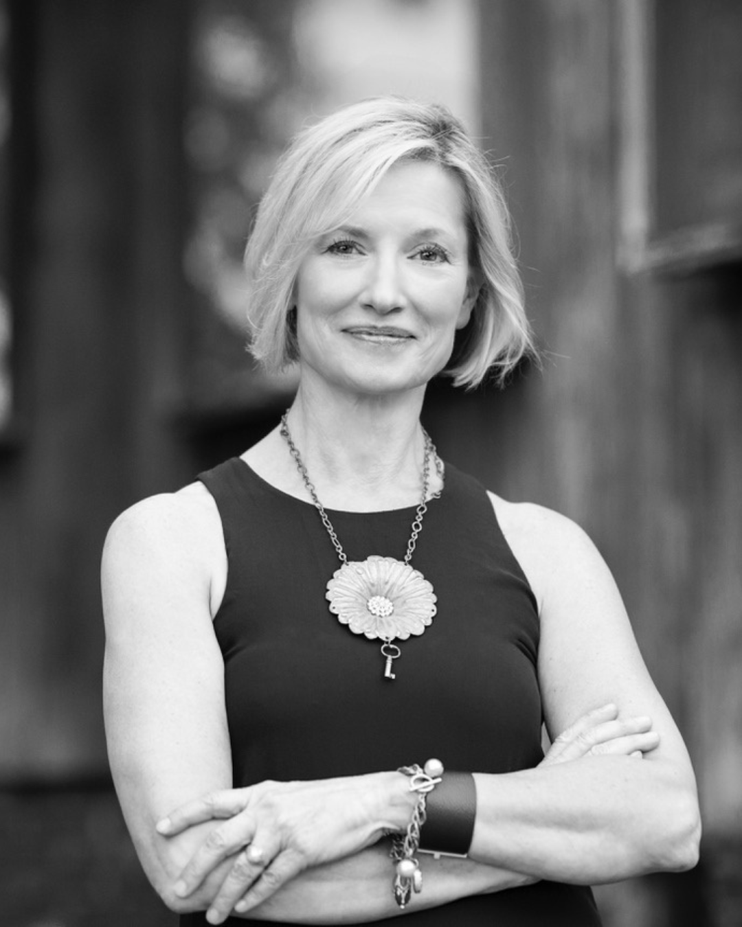 Black and white portrait of a woman with short blonde hair, wearing a sleeveless black top, with a large floral necklace, bracelets, and crossed arms, standing outdoors.
