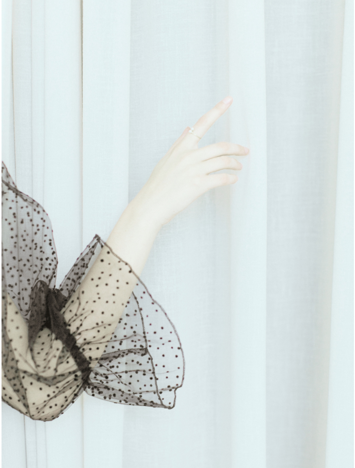Close-up of a woman's hand with a pearl ring, raised near a light-colored curtain, wearing a sheer polka-dotted sleeve with black trim.