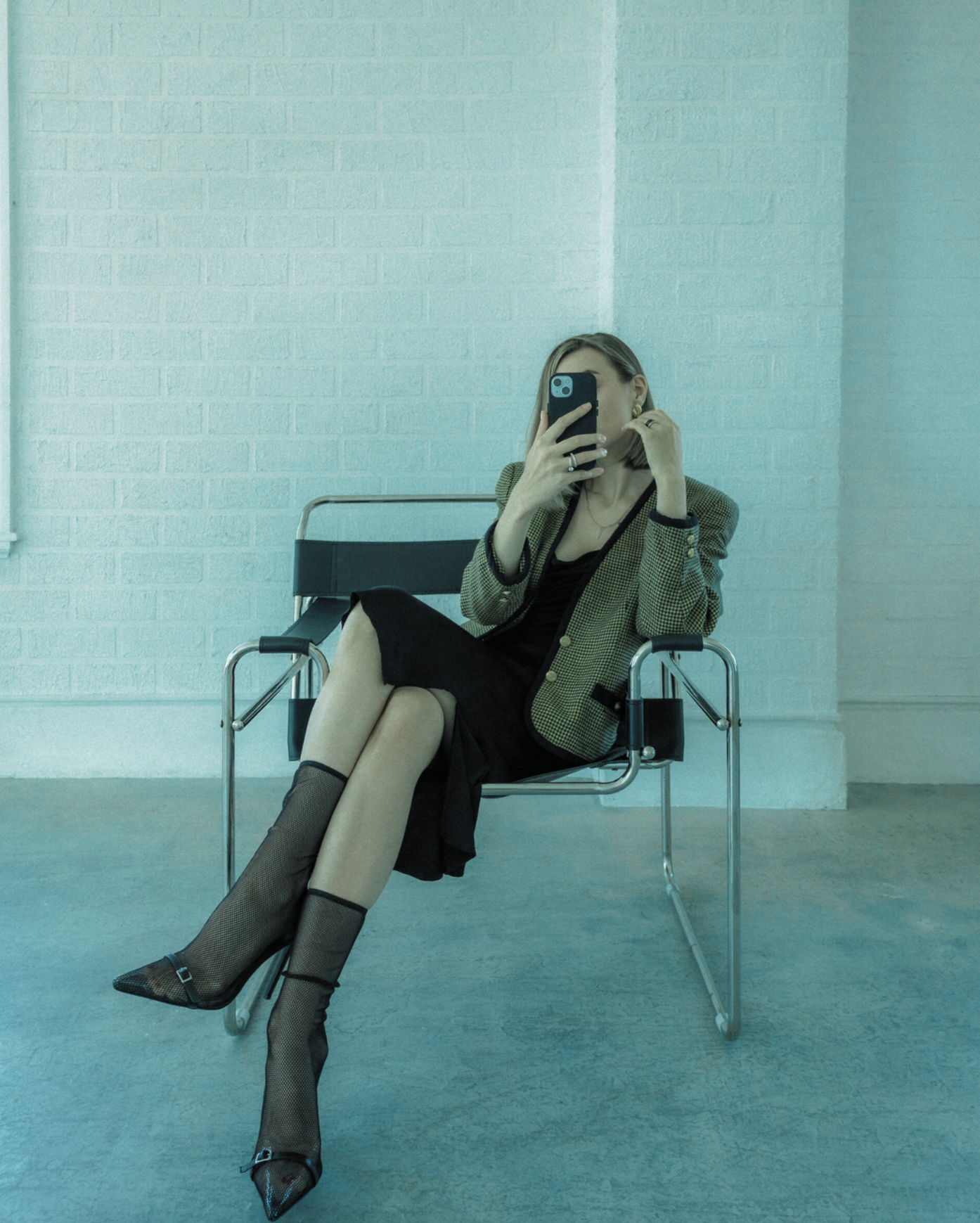 A woman sitting on a chair taking a selfie with her phone, wearing a black dress, fishnet stockings, pointed heels, and a checkered blazer.
