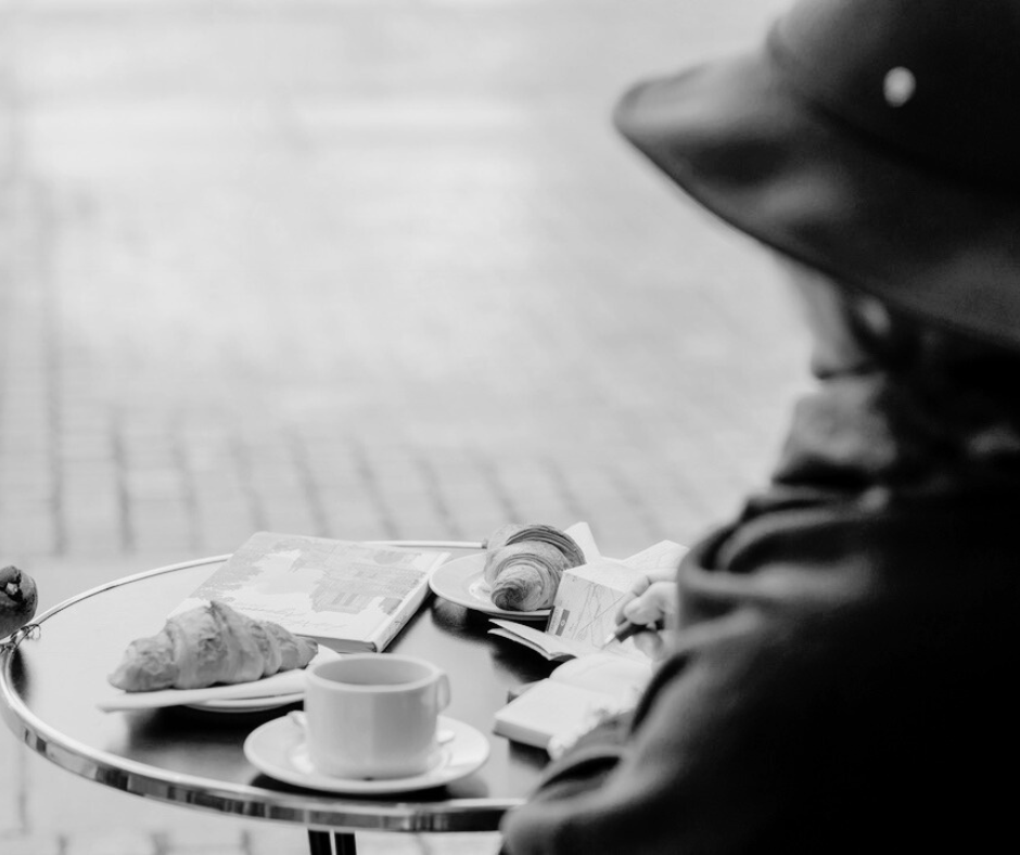 Person reading a newspaper and writing in a notebook at a small round table with a croissant, coffee cup, and a croissant pastry.