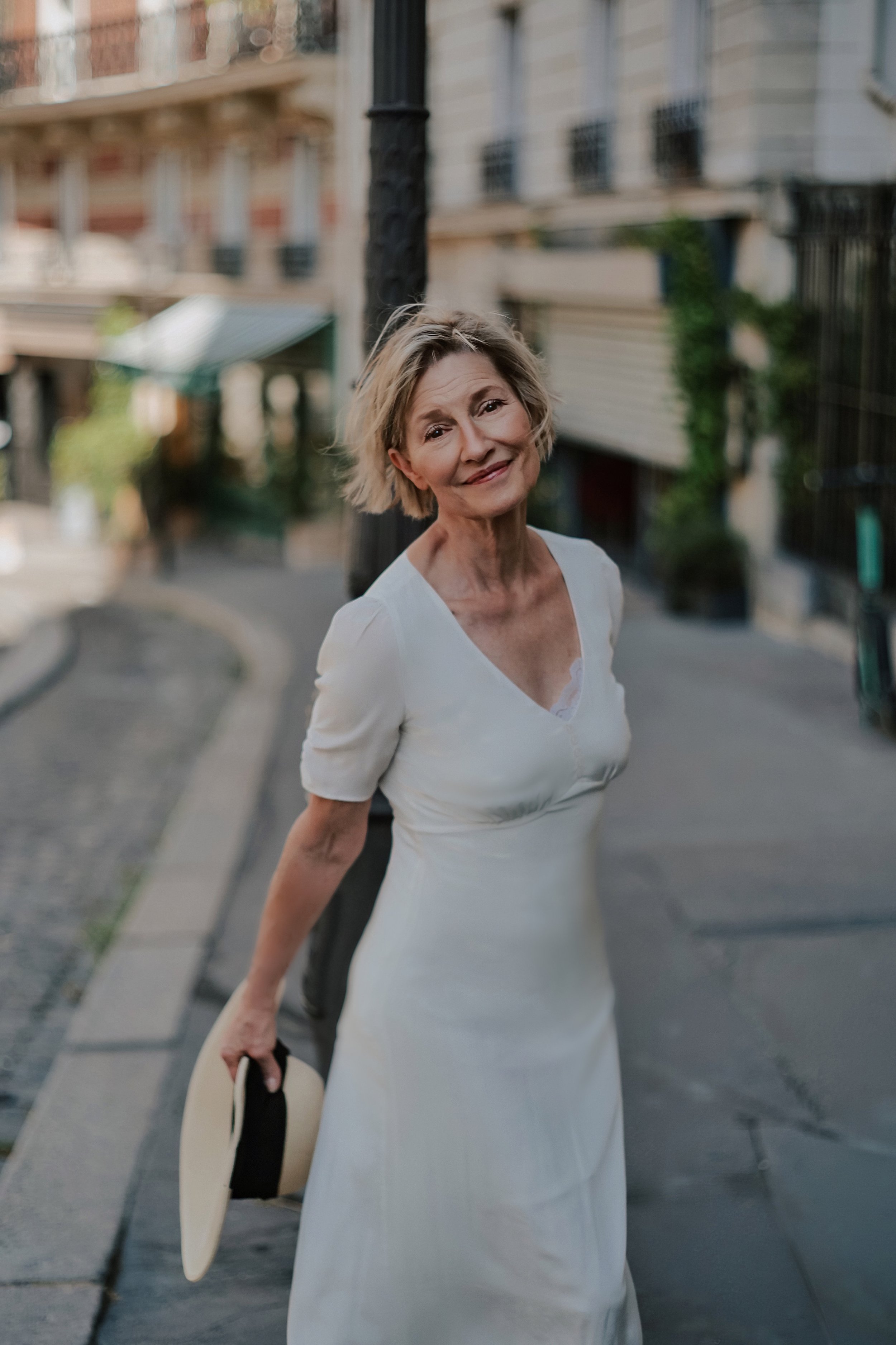 An older woman in a white dress holding a straw hat and standing on a city street, smiling at the camera.