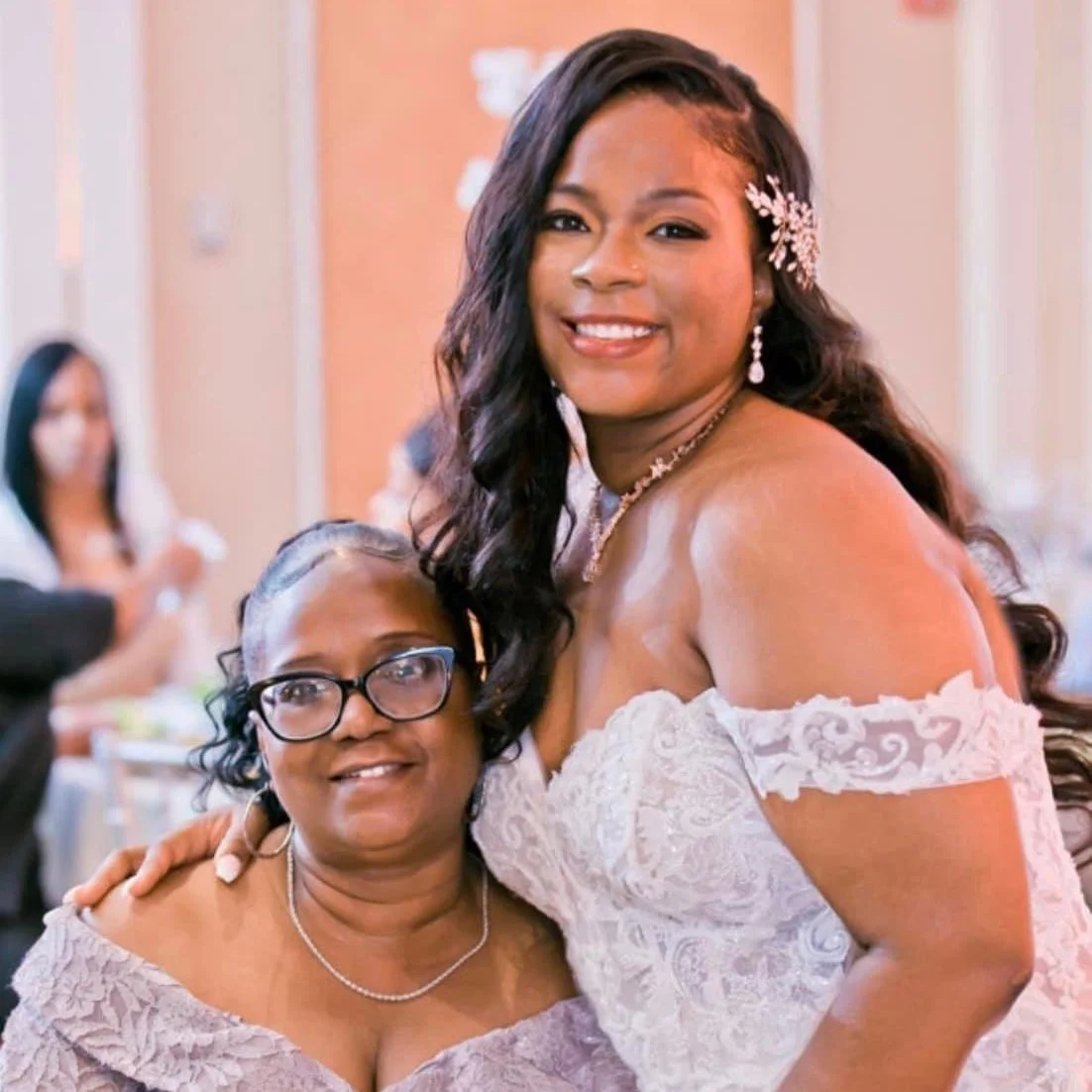 Two women dressed in wedding attire, one with long dark hair and the other with glasses, smiling at a wedding reception.