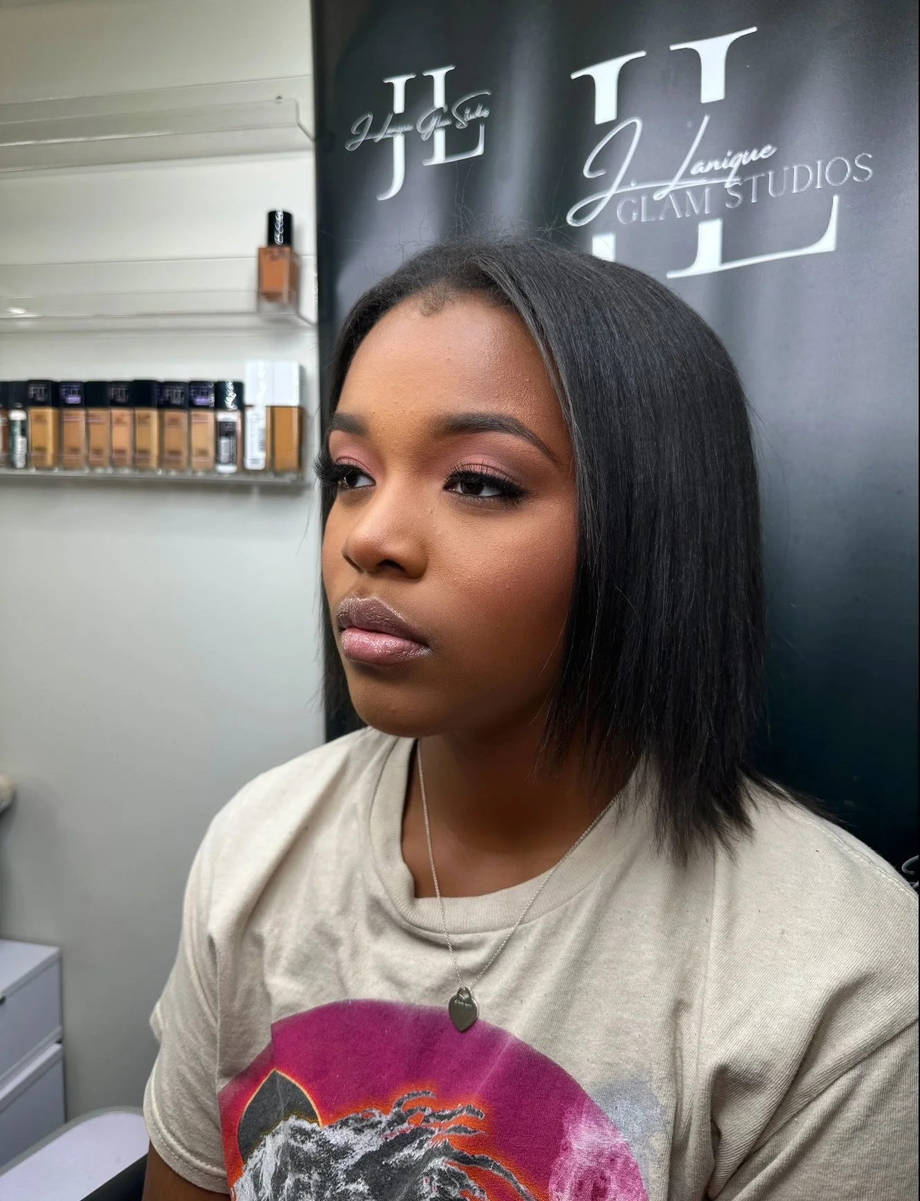 A young woman with straight black hair and makeup, wearing a beige T-shirt with a graphic design, and a silver necklace with a heart-shaped pendant, sitting indoors near a black wall with white text and shelves with makeup products.