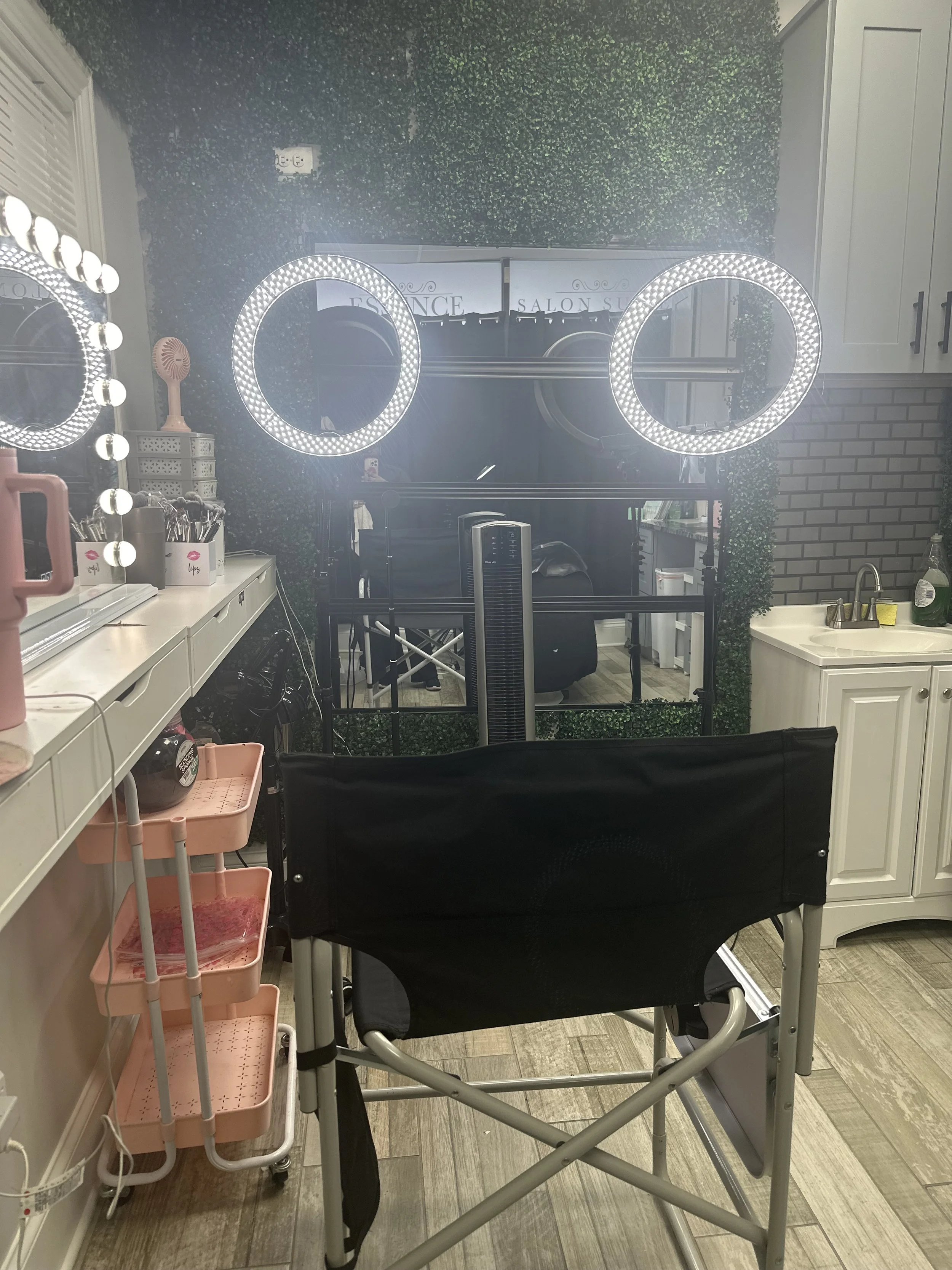 Beauty salon with illuminated ring lights, a mirror, makeup tools on the counter, and a black salon chair facing the mirror.