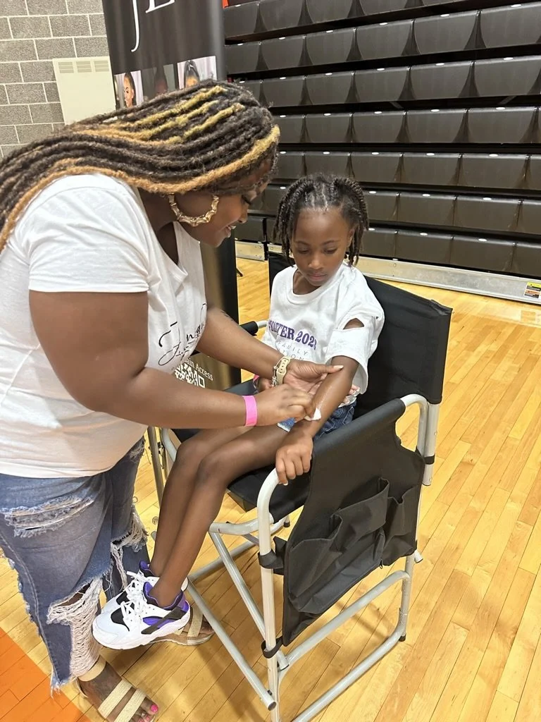 A woman is giving an injection to a young girl in a wheelchair. The girl appears calm, and the woman is smiling.