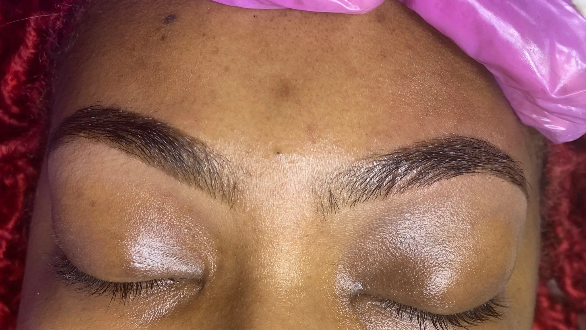 Close-up of a person's closed eyes and eyebrows, with freshly groomed eyebrows and smooth skin, lying on a pink fabric surface.