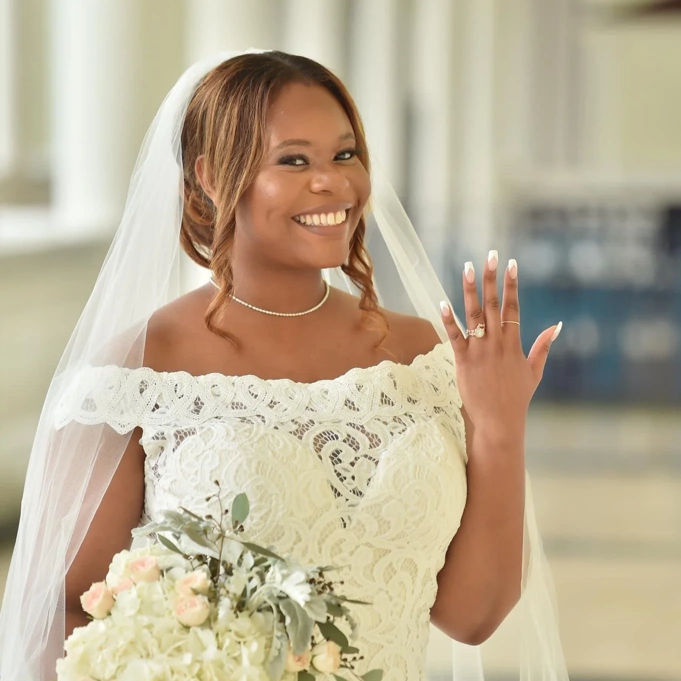 Woman in a wedding dress and veil showing her wedding ring, holding a bouquet of flowers, smiling happily.
