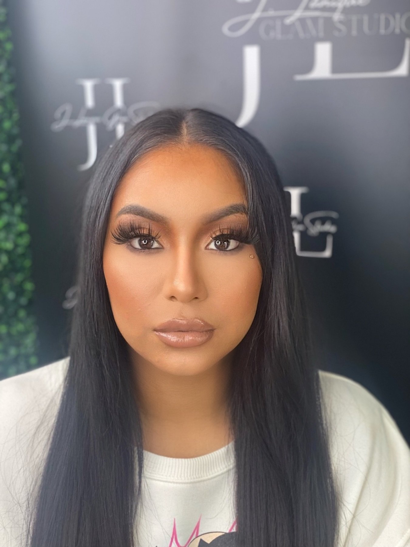 A woman with long black hair, makeup, and glossy lips, sitting against a black backdrop with silver lettering.