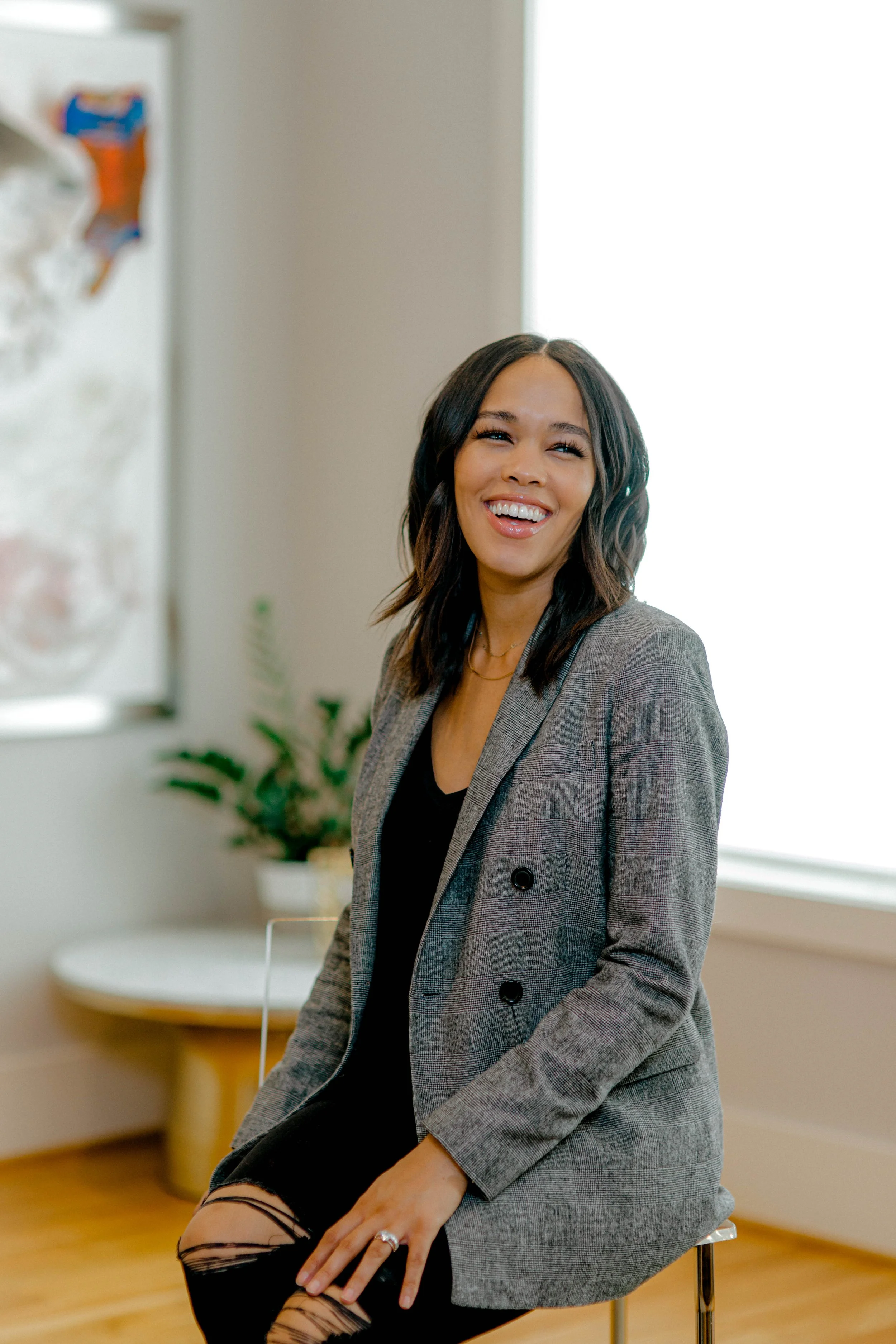 A woman with shoulder-length dark hair, wearing a gray blazer over a black top, sitting on a stool and smiling in a well-lit room with a window and a potted plant in the background.