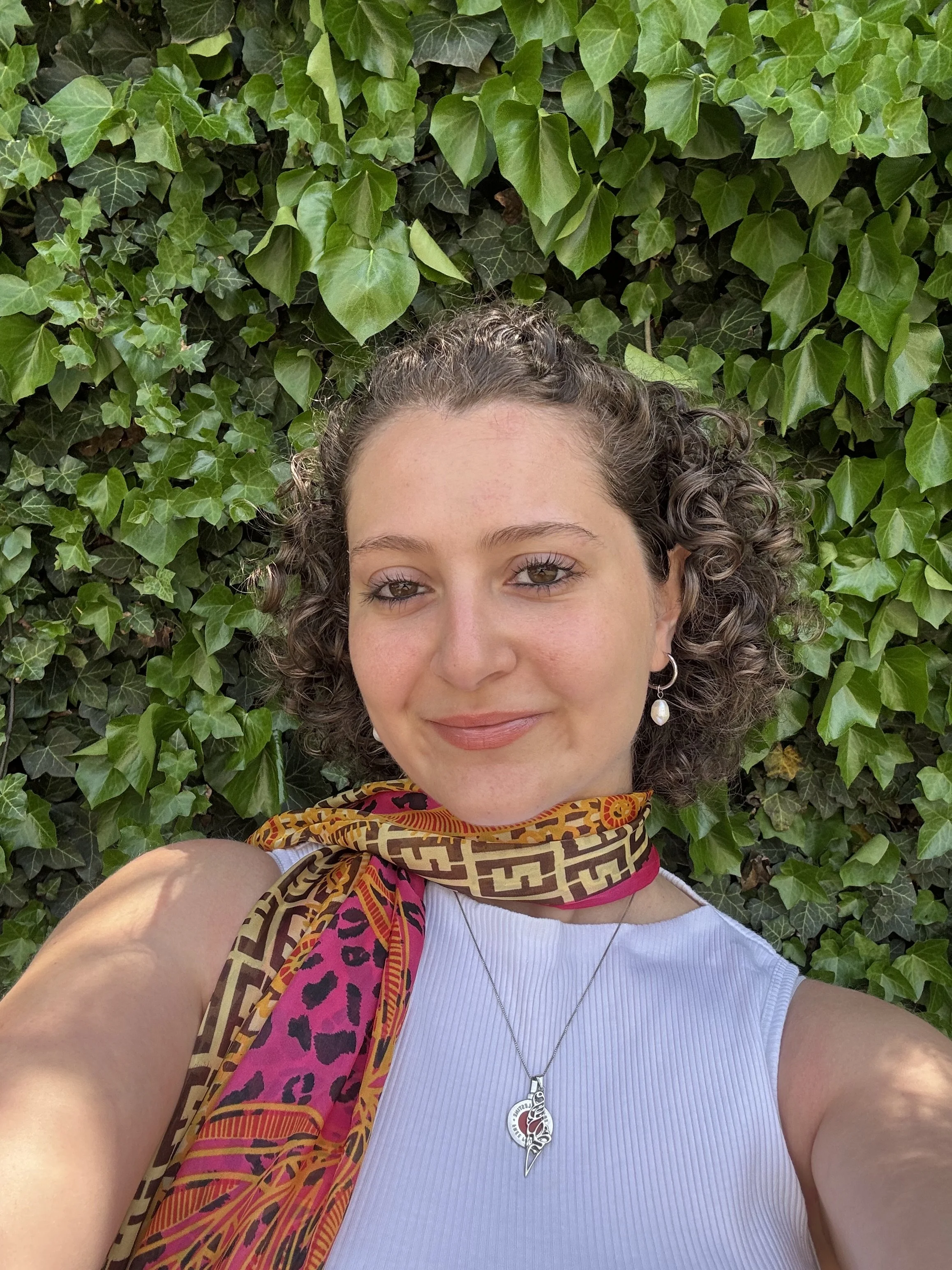 A woman with curly hair smiling and wearing a white sleeveless top, a colorful scarf, and a silver necklace with a pendant, taking a selfie in front of green ivy leaves.