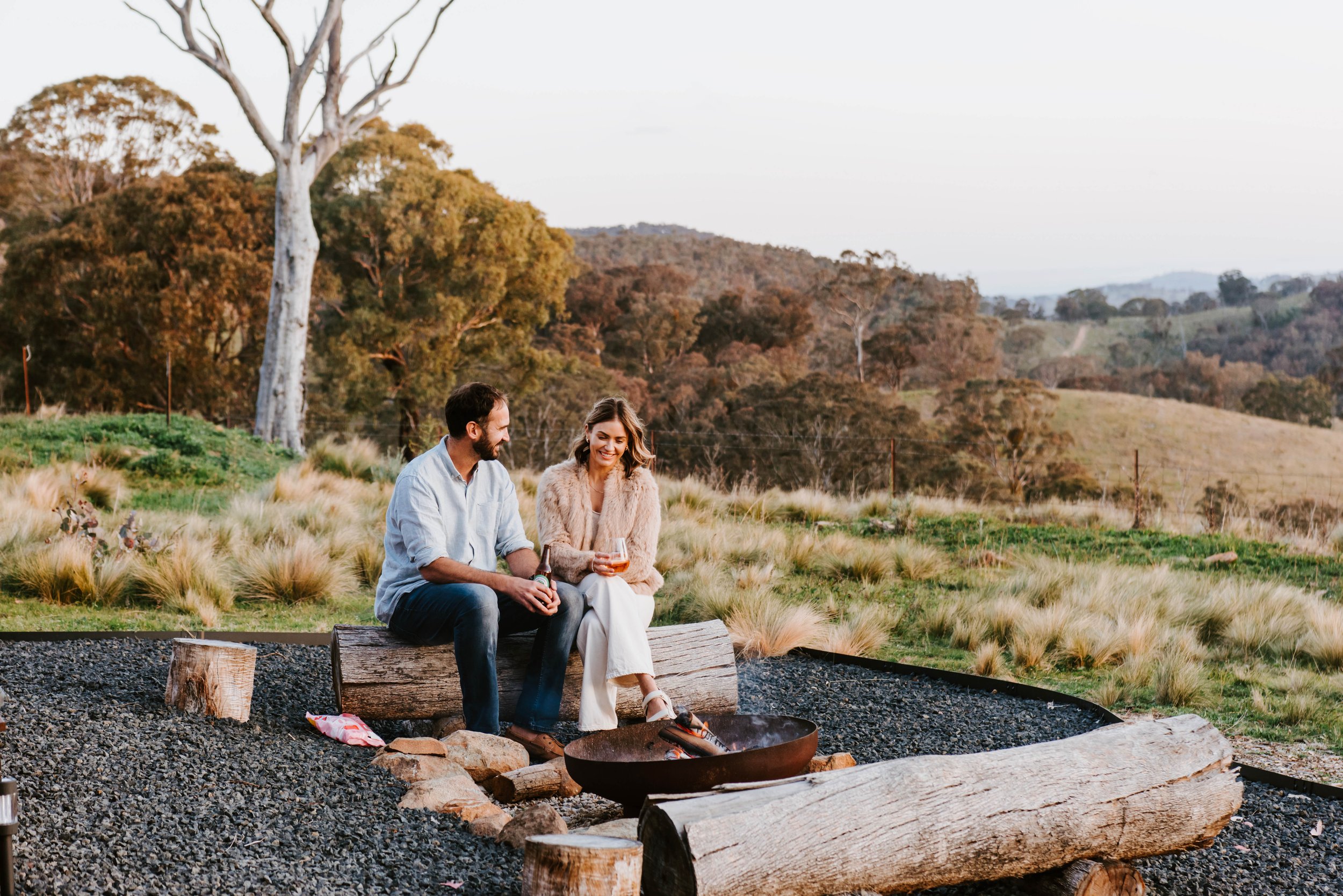A man and woman sitting on a wooden bench near an outdoor fire, smiling and enjoying drinks with a scenic landscape of trees and hills in the background during sunset.