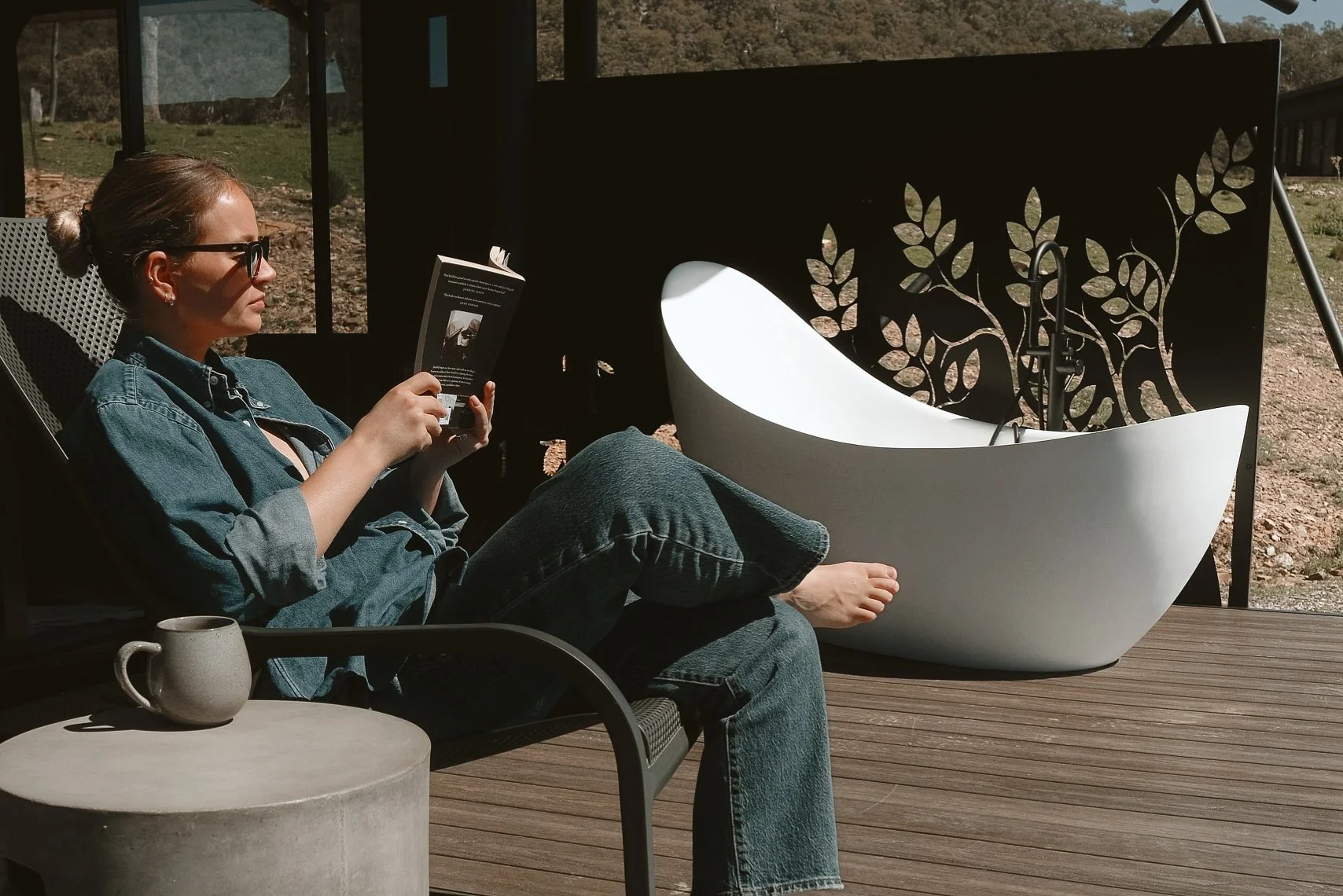 A guest reading a book with a cup of coffee on the deck of a glamping tent, with an outdoor bath overlooking the landscape at Wildnest Wilderness near Bathurst, NSW, photography by Helen Cooper