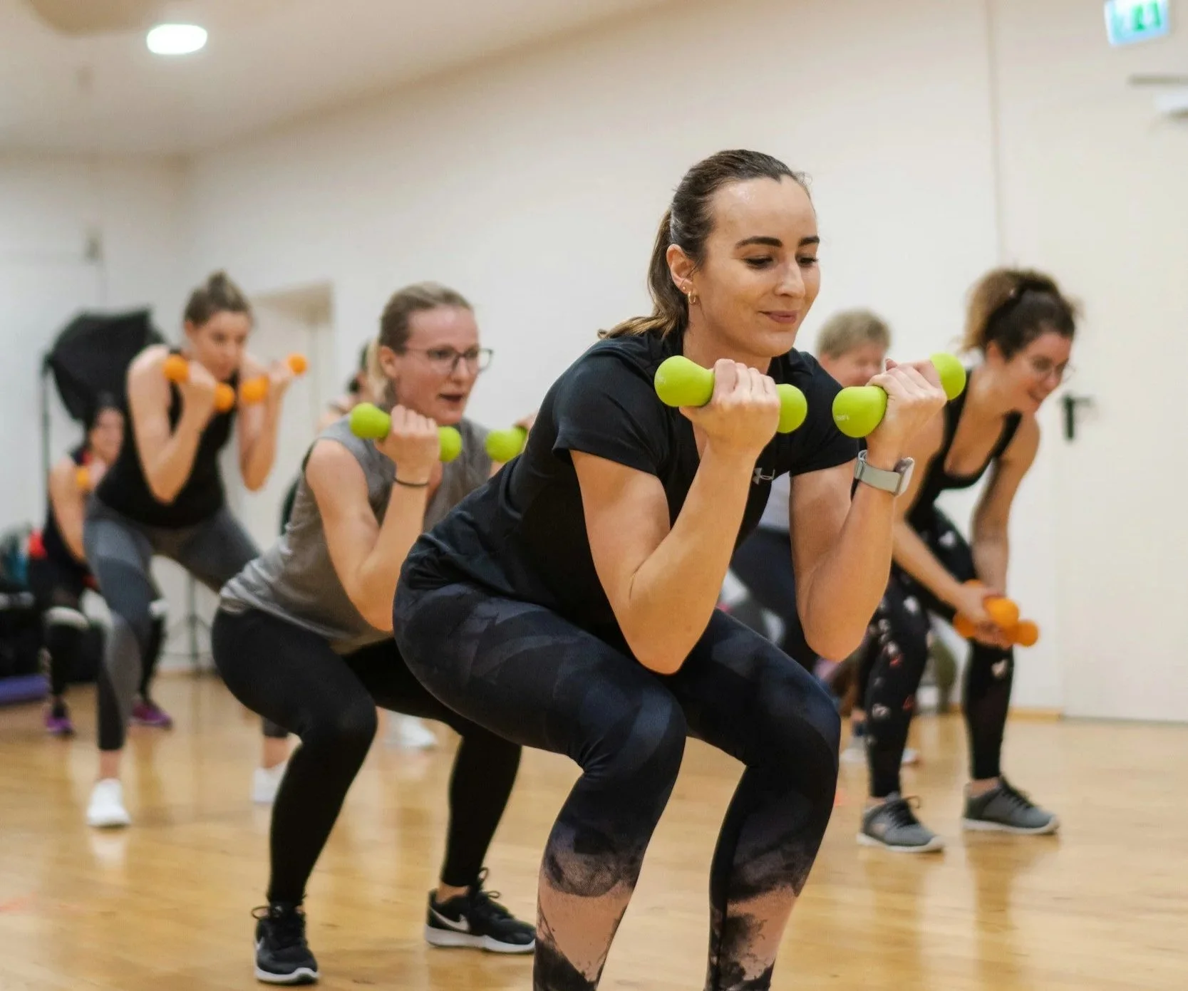 Women participating in a group indoor workout class, performing squats with small dumbbells, focused on exercise. The room has a wooden floor and plain walls.