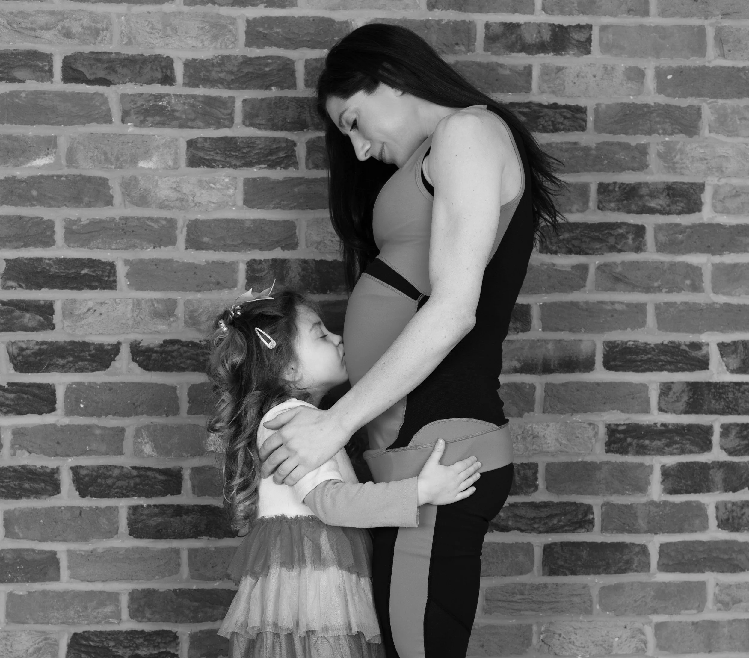 A woman in a black tank top and a girl in a tutu are hugging against a brick wall. The woman is looking down at the girl, who has curly hair decorated with clips, and they are embracing each other.