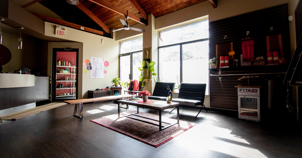 Bright interior of a cozy waiting or lounge area with large windows, black chairs, a coffee table, plants, and decorative items.