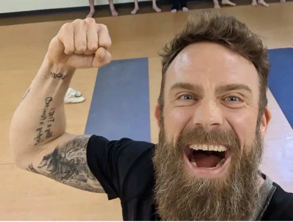 Happy man with a beard taking a selfie in a gym or yoga studio, flexing his arm with tattoos, and smiling broadly, with other people in the background.