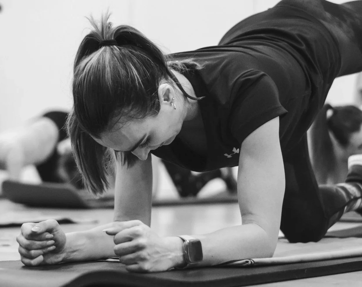 A woman performs a plank exercise on a yoga mat in a fitness class, with other participants visible in the background.