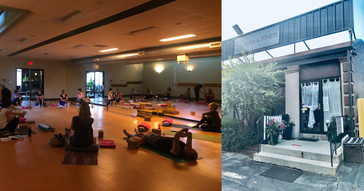 A yoga class taking place inside a spacious studio with wooden floors, where participants are seated on yoga mats, engaging in a pose and guided by an instructor. The studio has a mirrored wall and large windows bringing in natural light. Next to it is an outdoor entrance to a yoga and fitness studio, with a sign overhead, surrounded by greenery and potted plants.