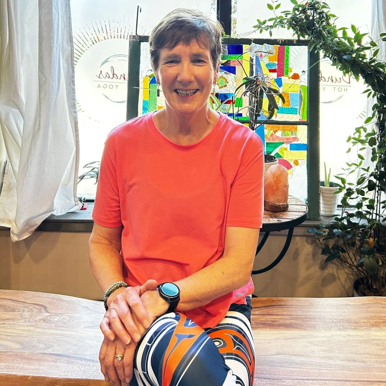 A woman with short brown hair smiling, wearing a coral pink T-shirt and striped leggings, sitting at a wooden table in a bright room with plants and a colorful stained glass window in the background.