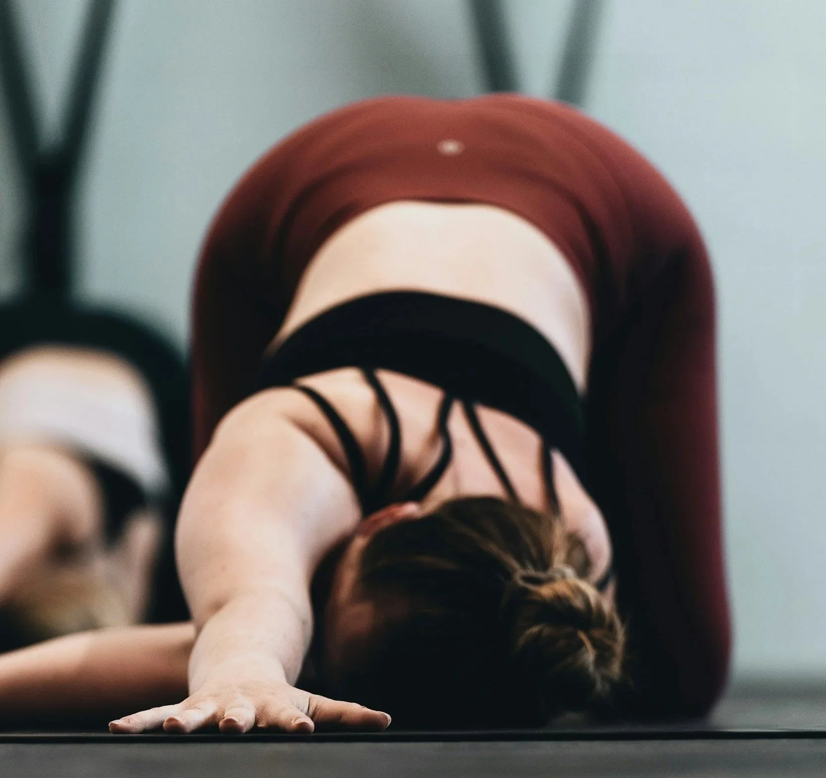 A person is doing a yoga pose on the floor with one arm extended forward and the head lowered to the ground, wearing workout clothes including a black top and patterned leggings, in a room with a plain background.