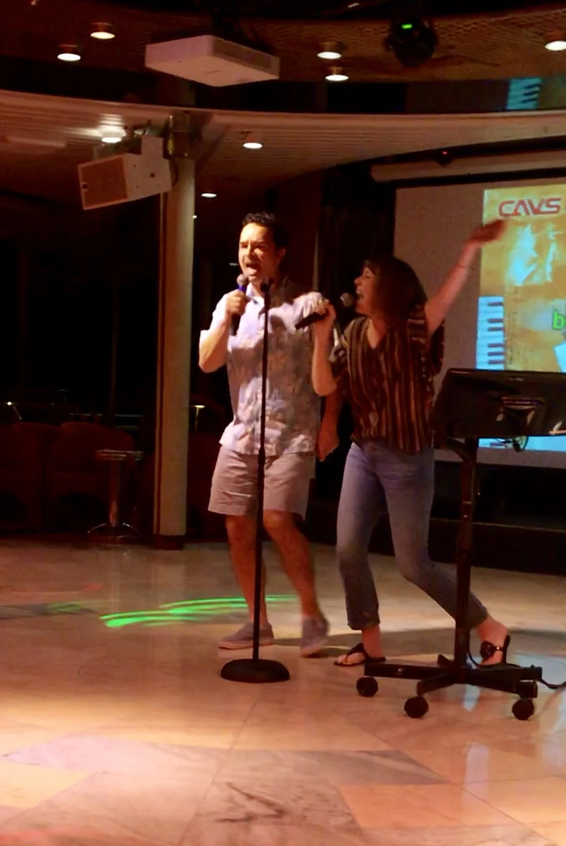 Two people singing karaoke on a stage with microphones, one male wearing shorts and a floral shirt, the other female in a striped blouse and jeans, in a dimly lit room with a screen in the background.