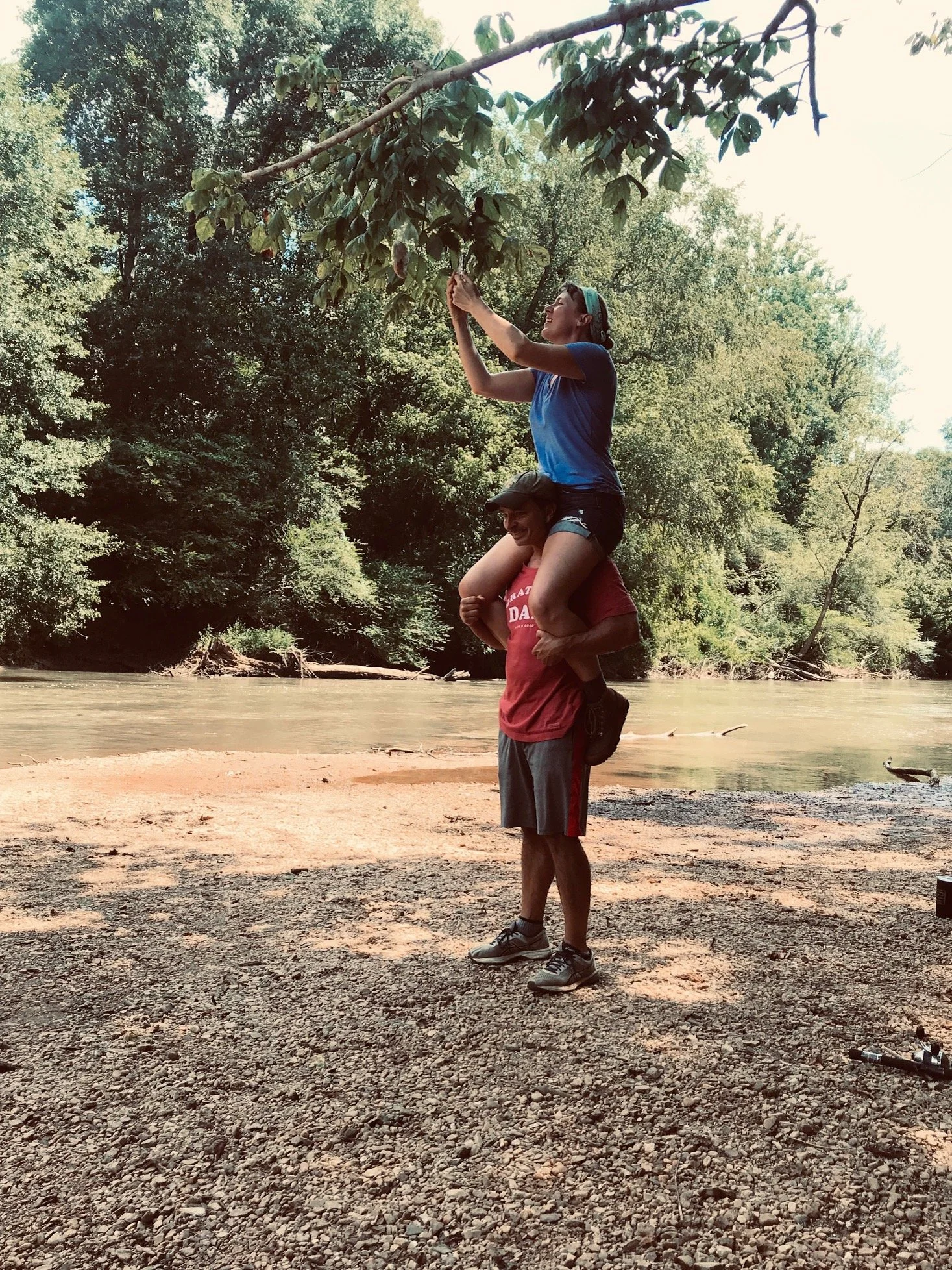 Two people, one standing and one sitting on the other person's shoulders, in a forested riverside area. The person on top is grabbing a tree branch, both smiling, surrounded by trees and water.