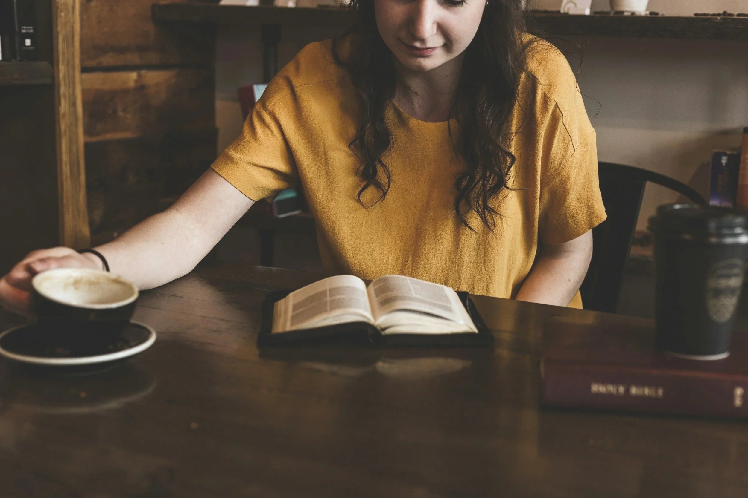 A woman with long dark hair wearing a mustard yellow shirt sitting at a wooden table, reading a book with an open pages. Several cups of coffee or tea are placed on the table. The background shows a bookshelf and a dark wall.
