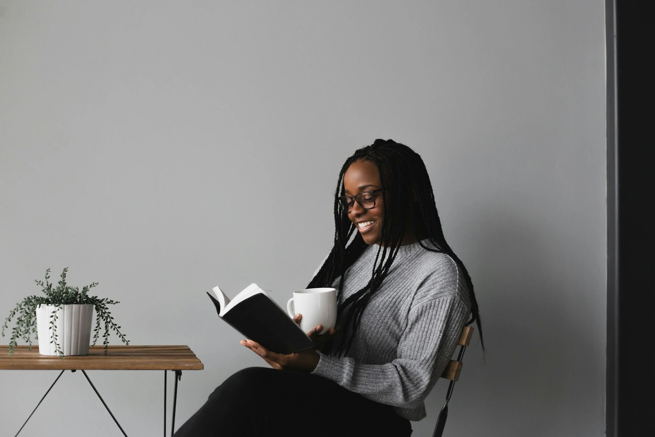 Person reading a book and holding a mug, sitting near a wooden table with a potted plant, set against a plain gray background.
