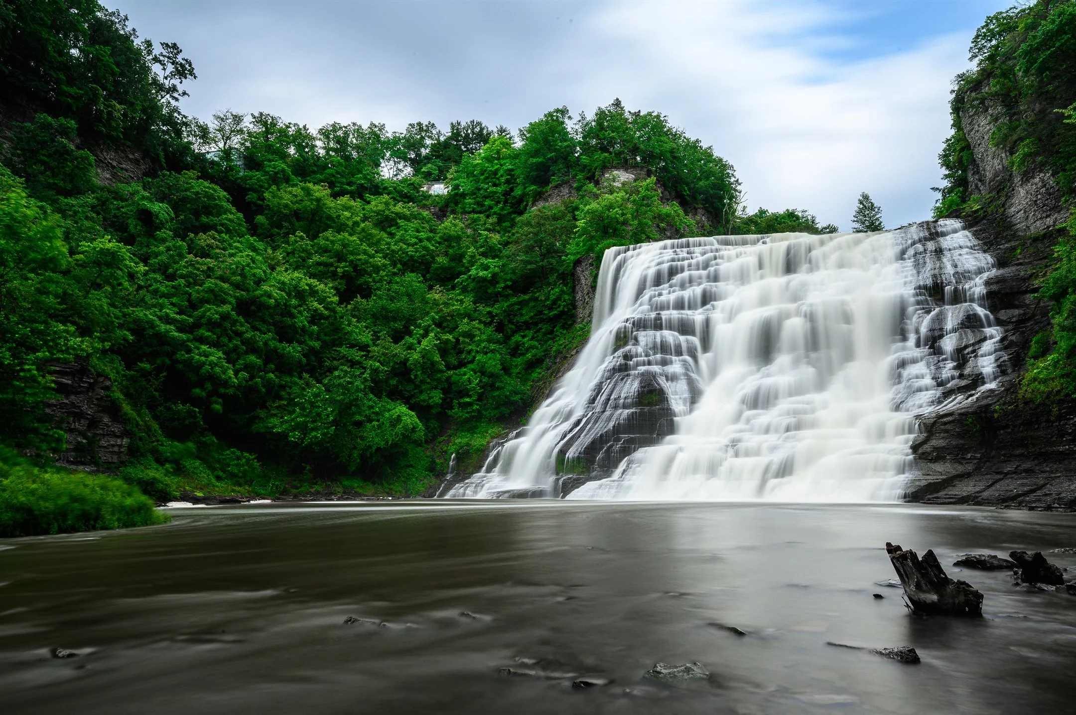 Waterfall with cascading water surrounded by lush green trees and rocky cliffs.