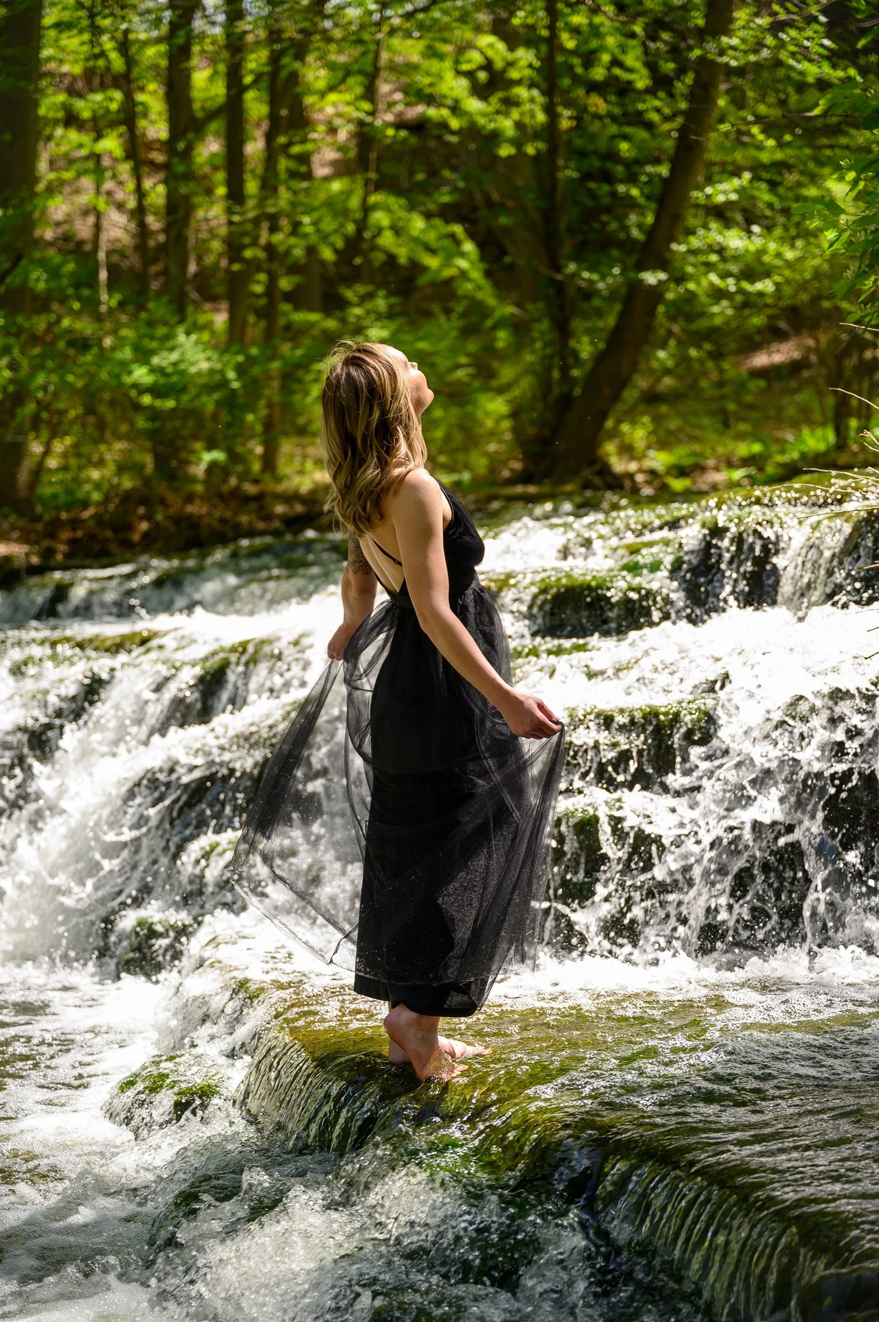 Woman in black dress standing on a small waterfall in a forest setting.