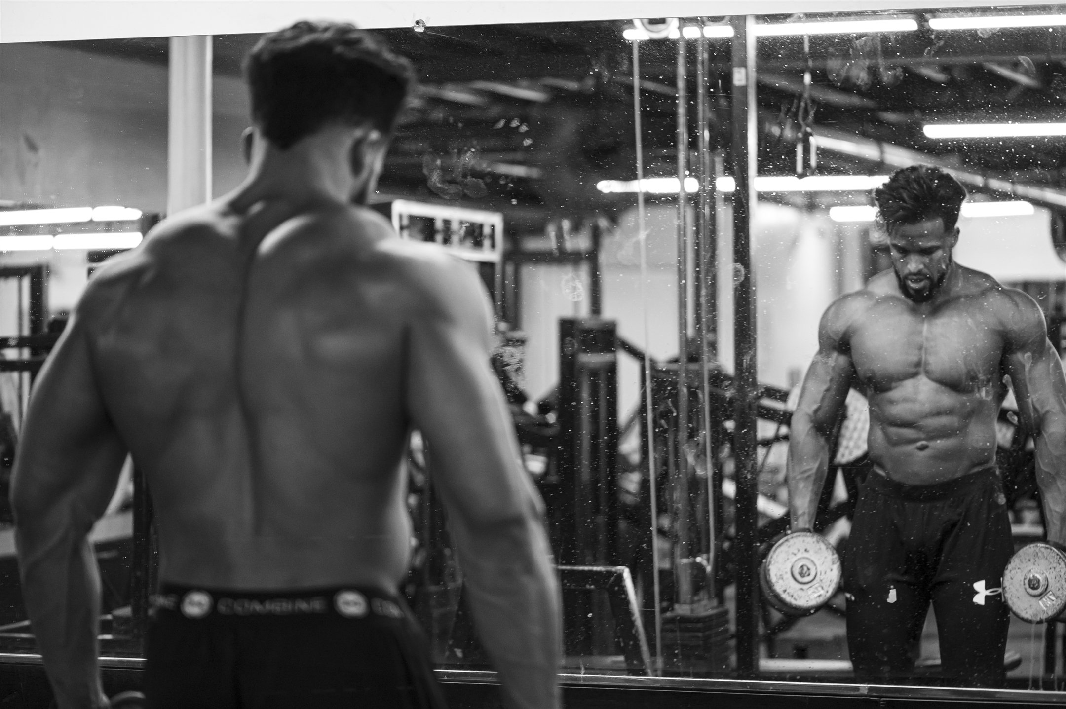 Bodybuilder lifting dumbbells in front of a mirror in a gym, black and white photo.