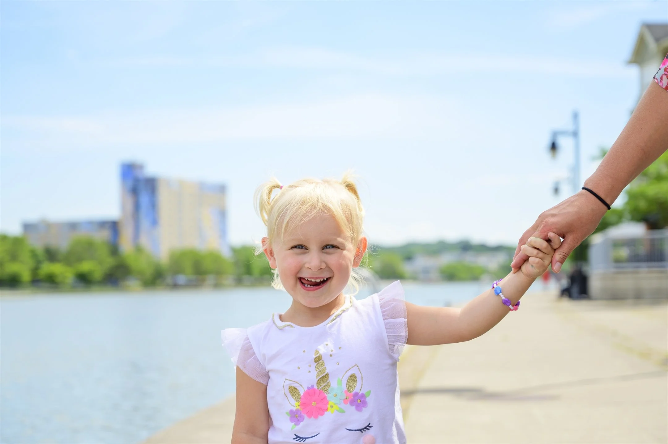 Smiling child in unicorn shirt holding adult's hand near waterfront.