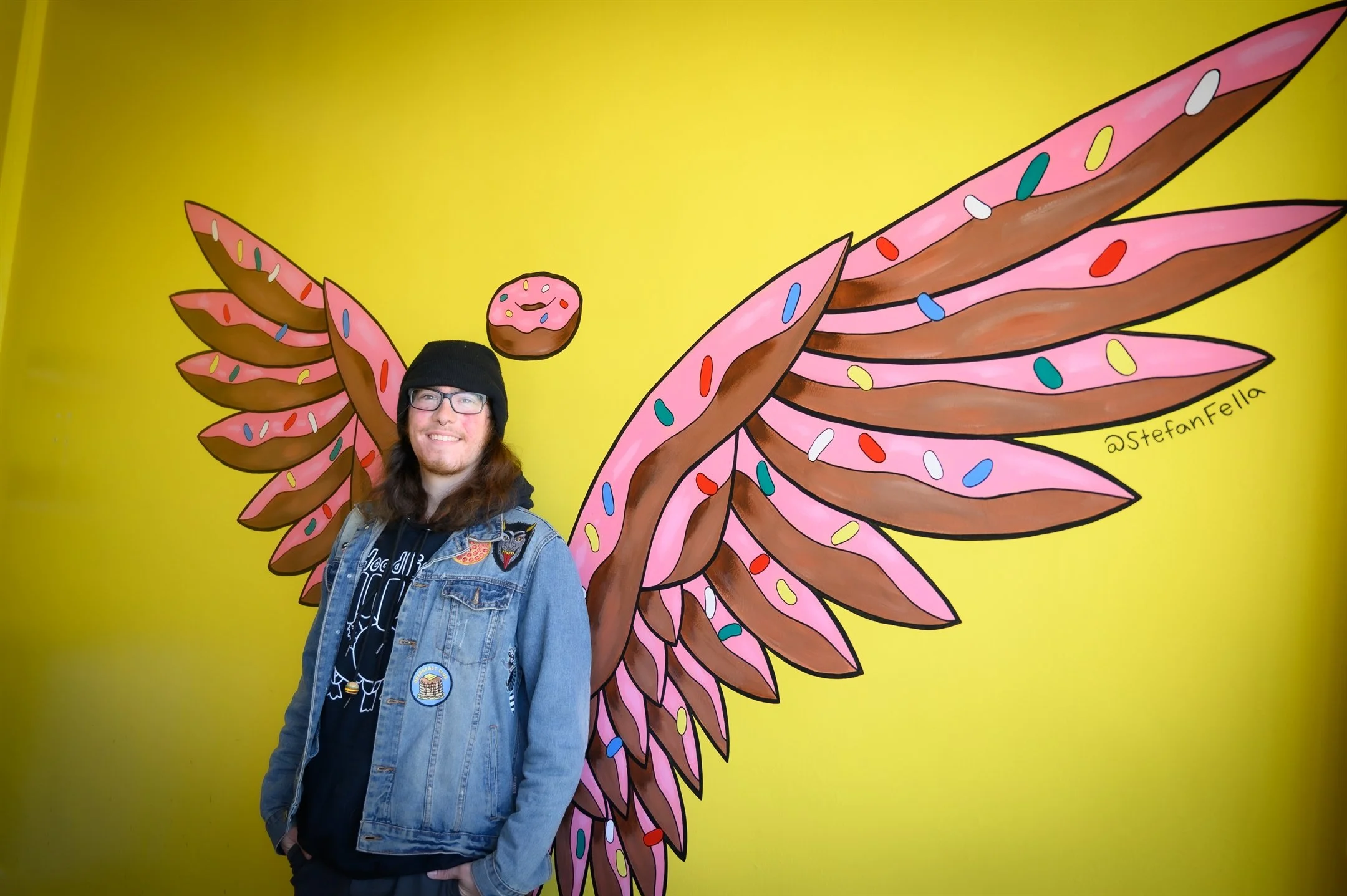 Person in a denim jacket and beanie standing in front of a yellow wall with a mural of donut wings and a small donut above their head.