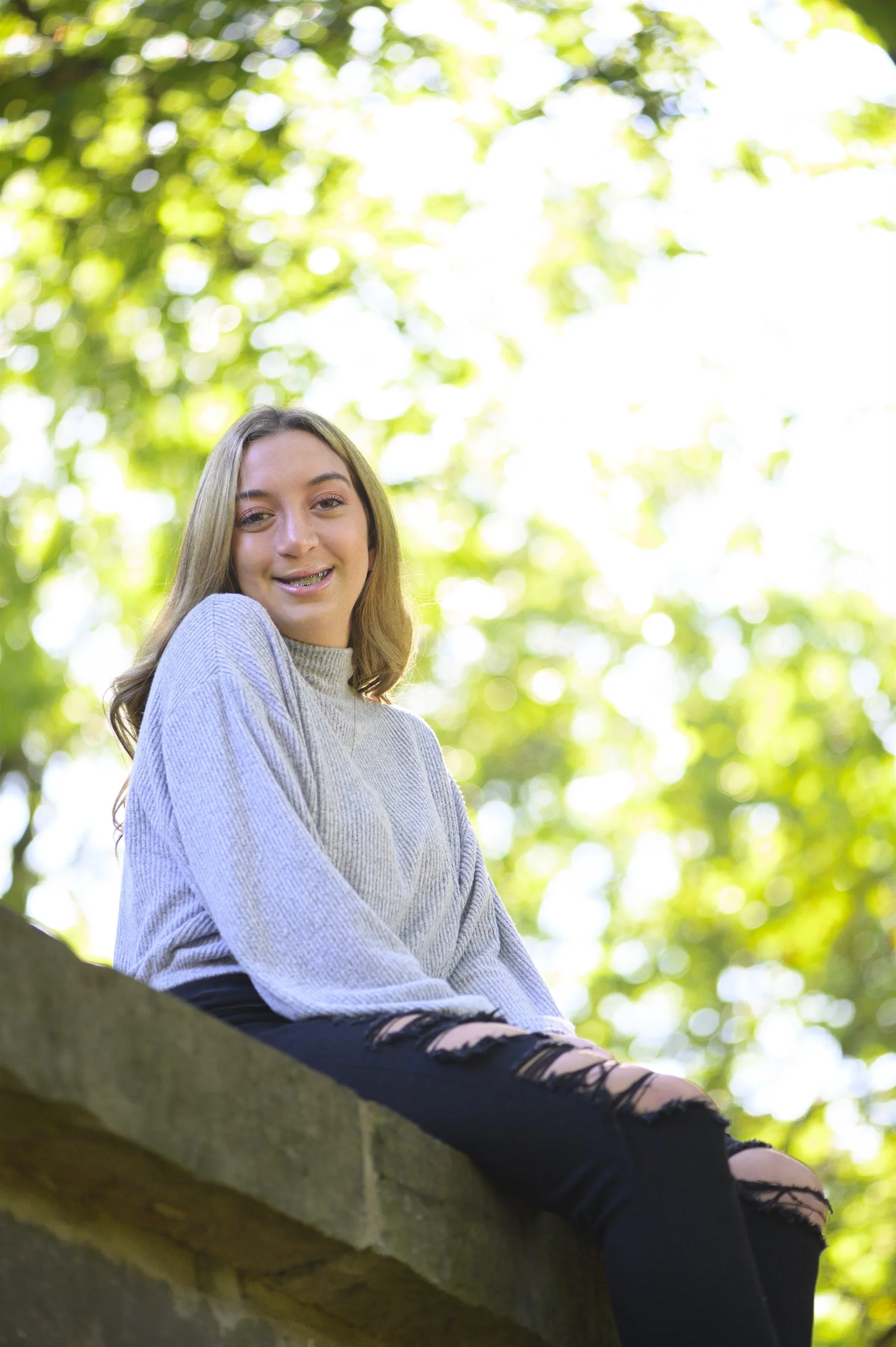 Smiling person sitting outdoors wearing a gray sweater and ripped jeans, with a blurred green leafy background.
