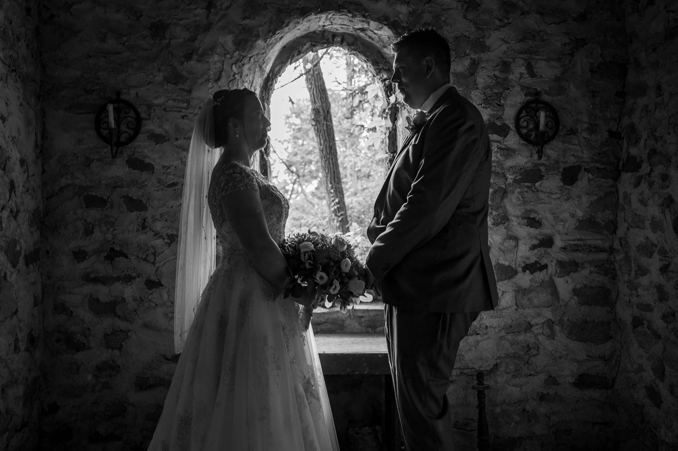 Silhouetted couple in wedding attire holding hands in front of an arched window with stone walls