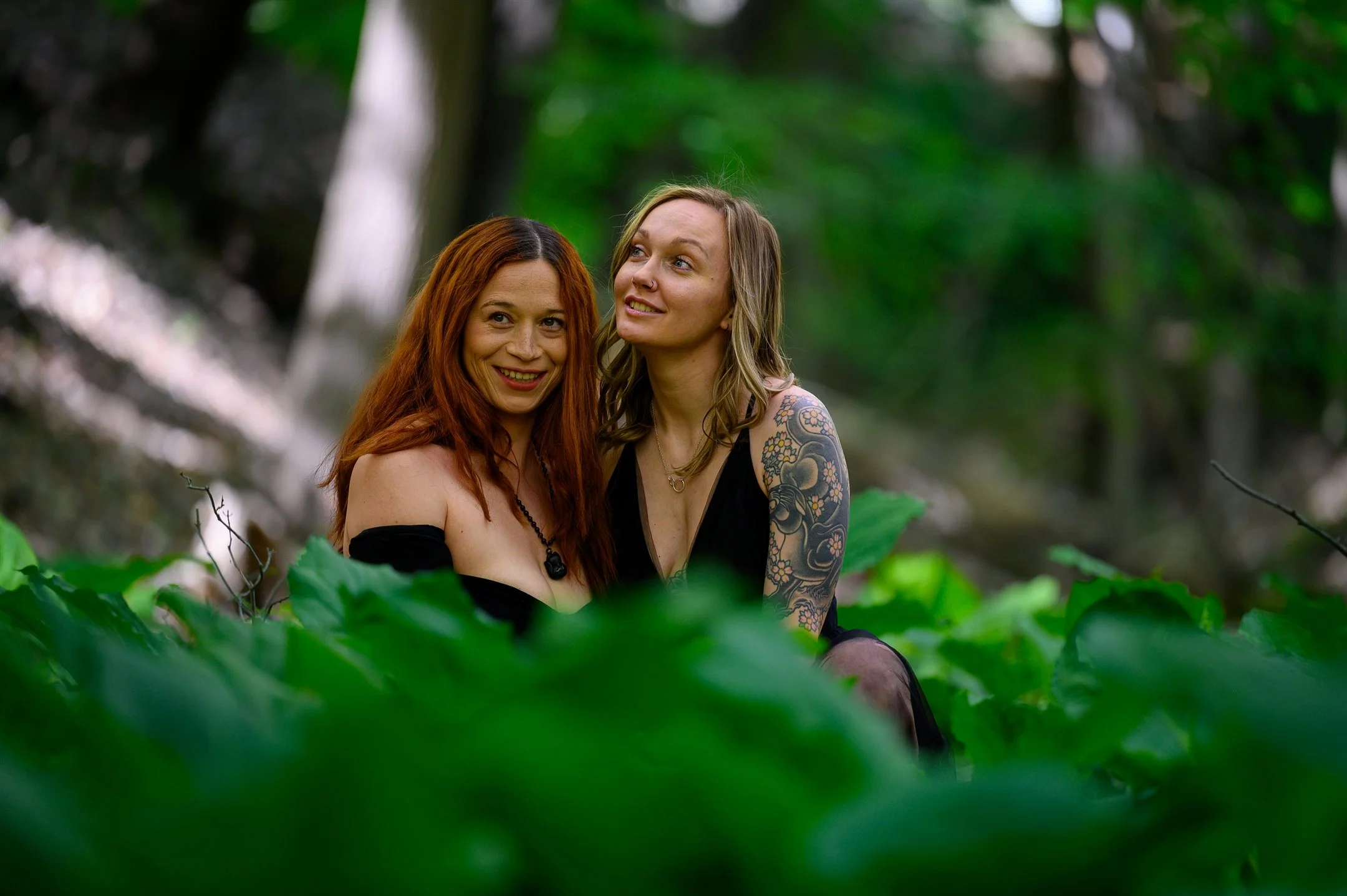 Two women sitting among green leaves in a forest, one with red hair and one with tattoos, both wearing black outfits.