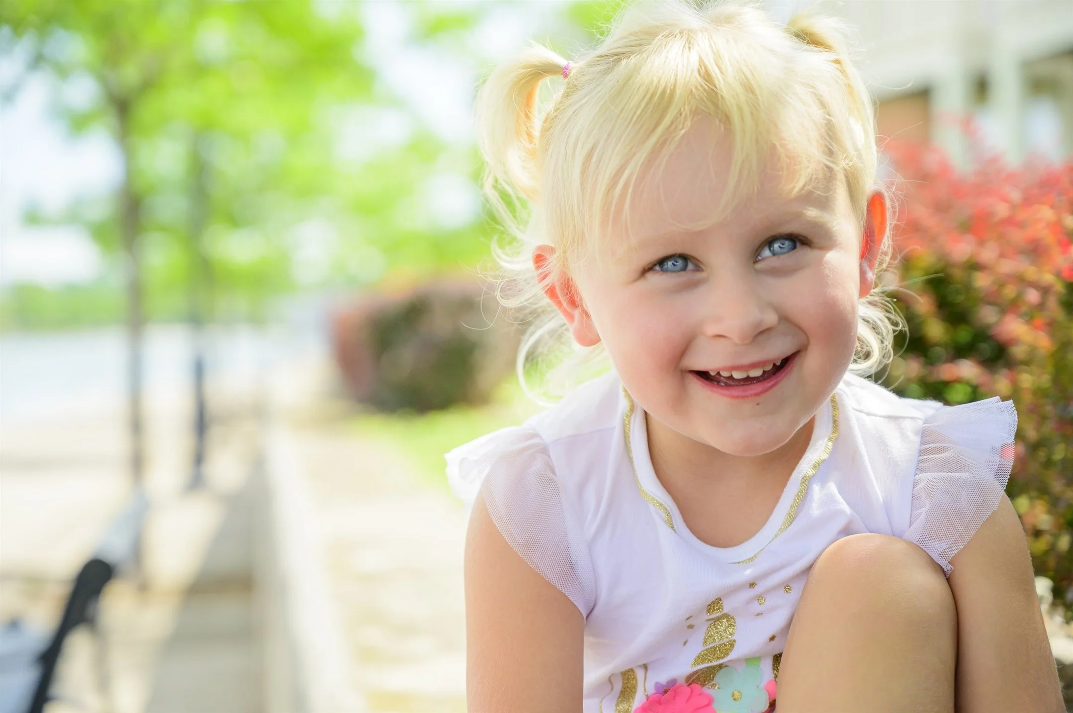 Smiling child with blonde hair and ponytails sitting outside on a sunny day, wearing a white shirt with colorful designs, with trees and blurred background.