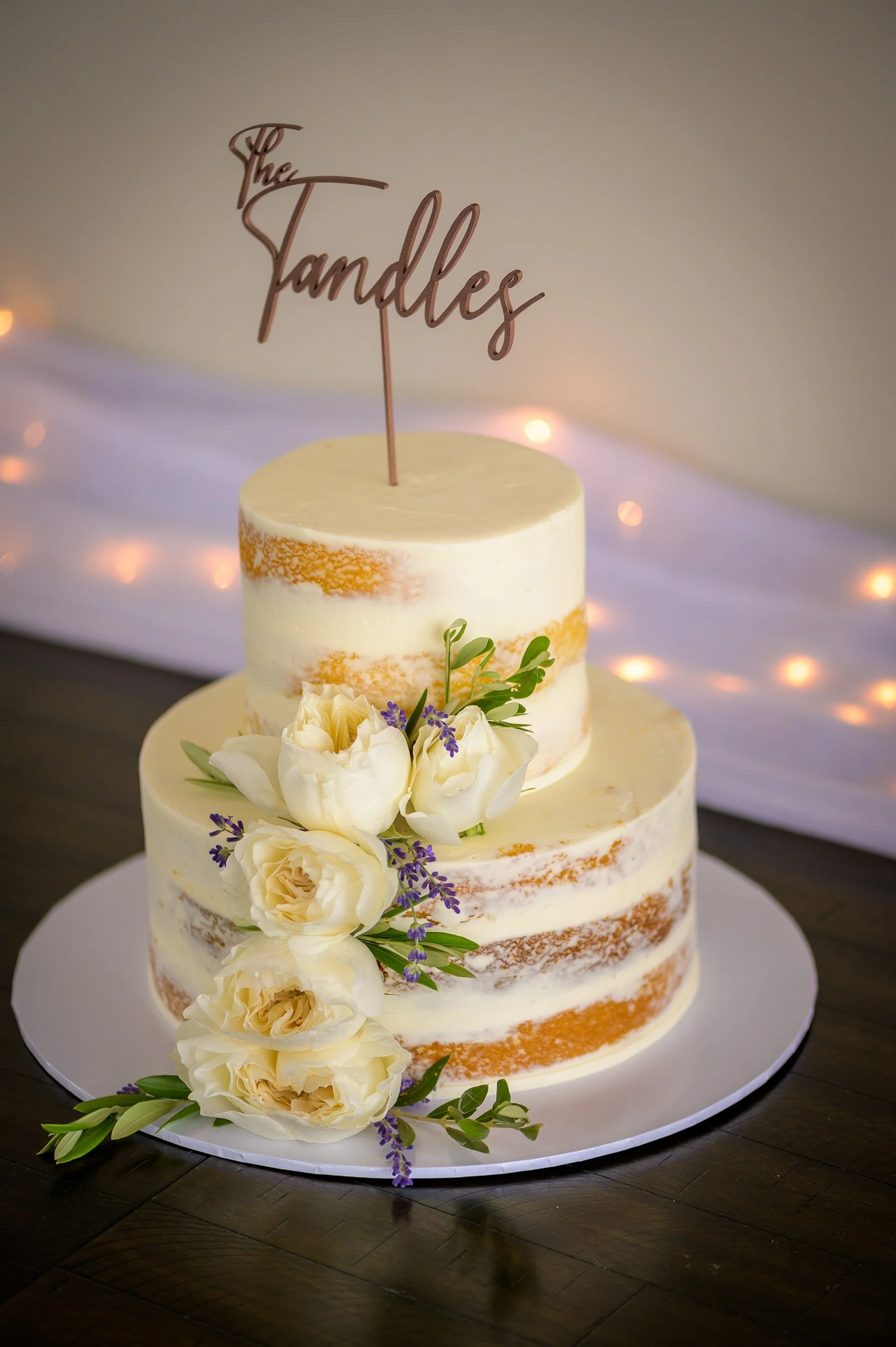 Two-tier naked wedding cake with a cake topper, decorated with white roses and lavender on top.