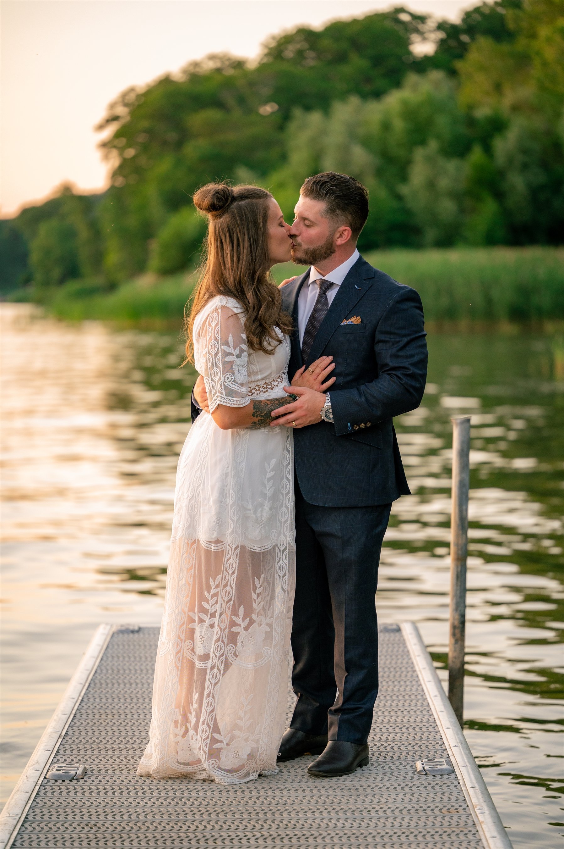 A couple kissing on a dock by a lake, surrounded by greenery, during sunset. The woman is wearing a white lace dress, and the man is in a dark suit.