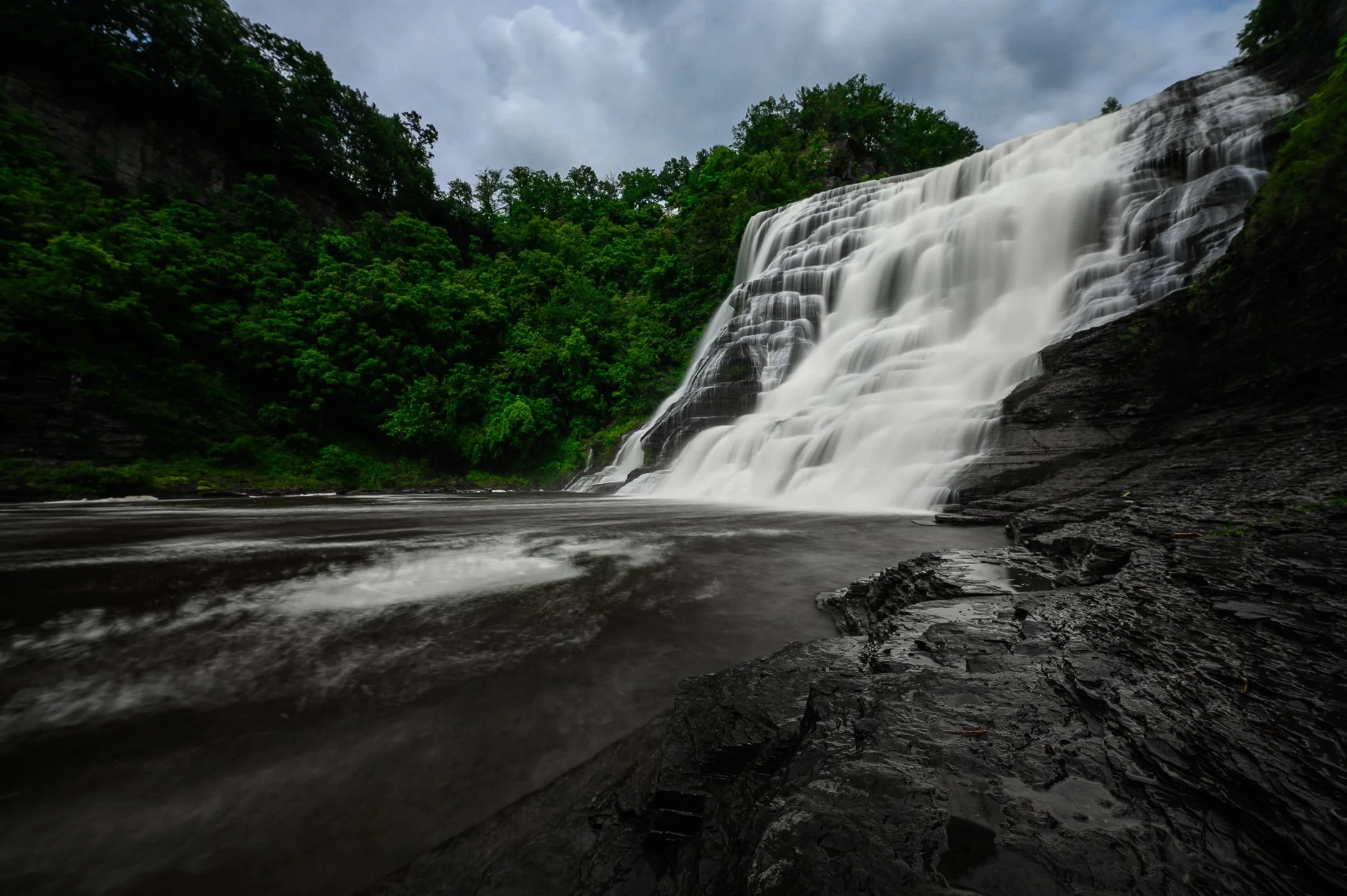 Waterfall cascading over rocky cliffs into a calm river, surrounded by lush green foliage under a cloudy sky.