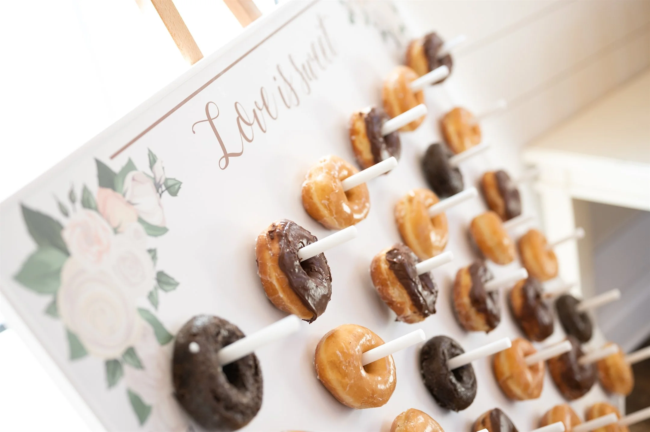 A wedding donut wall display with glazed and chocolate donuts on sticks, decorated with floral designs and a "Love is sweet" sign.