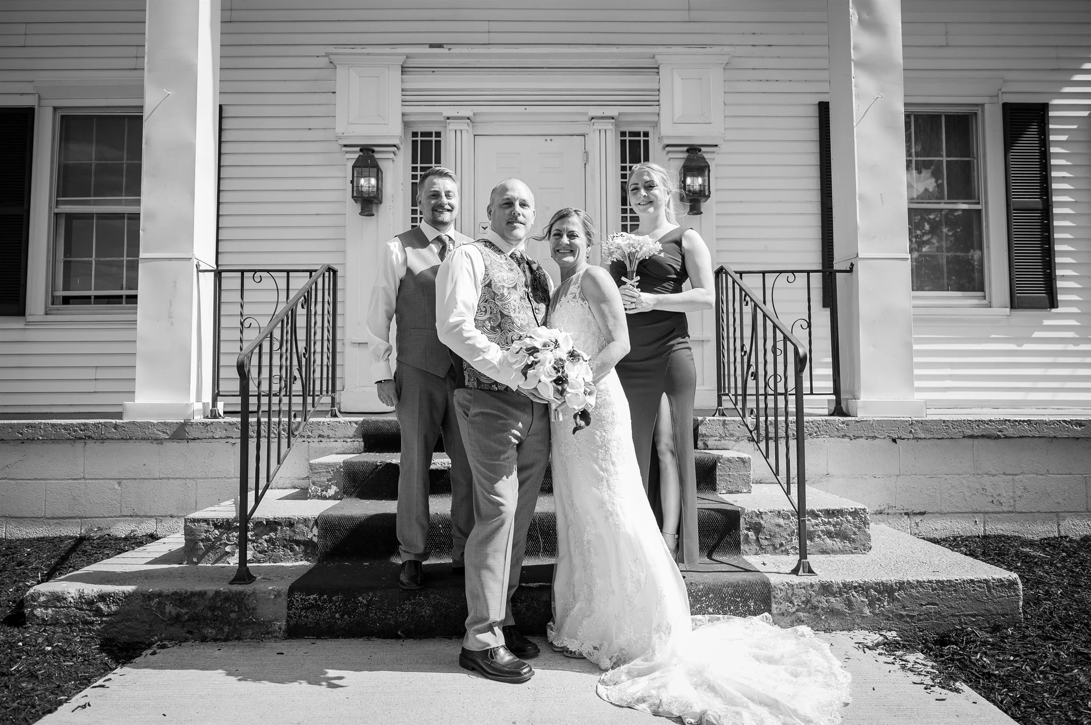Black and white photo of a wedding party posing in front of a building with white siding and a staircase. The group includes the bride in a lace gown, groom in a vest and tie, and two attendants, one holding a bouquet.