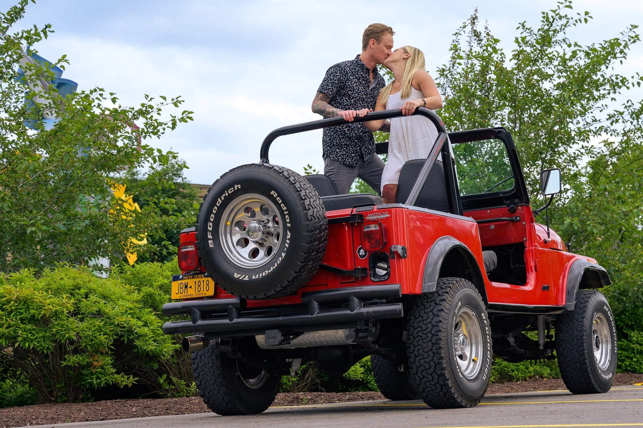 A couple kissing while standing in the back of a red convertible Jeep parked outdoors, surrounded by greenery and under a cloudy sky.